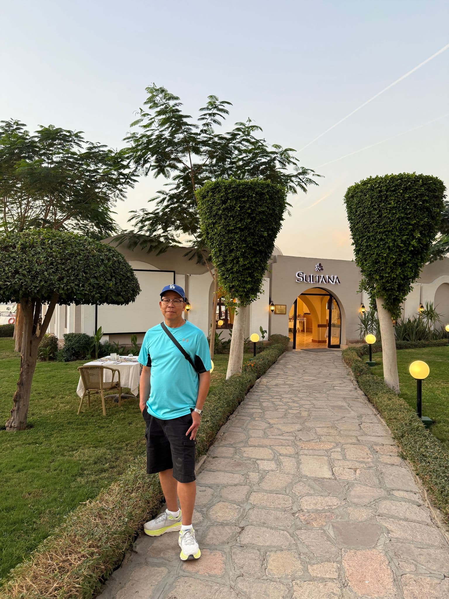 Sultana Hotel entrance in Luxor, Egypt, with a traveler standing on a stone path flanked by trimmed trees.