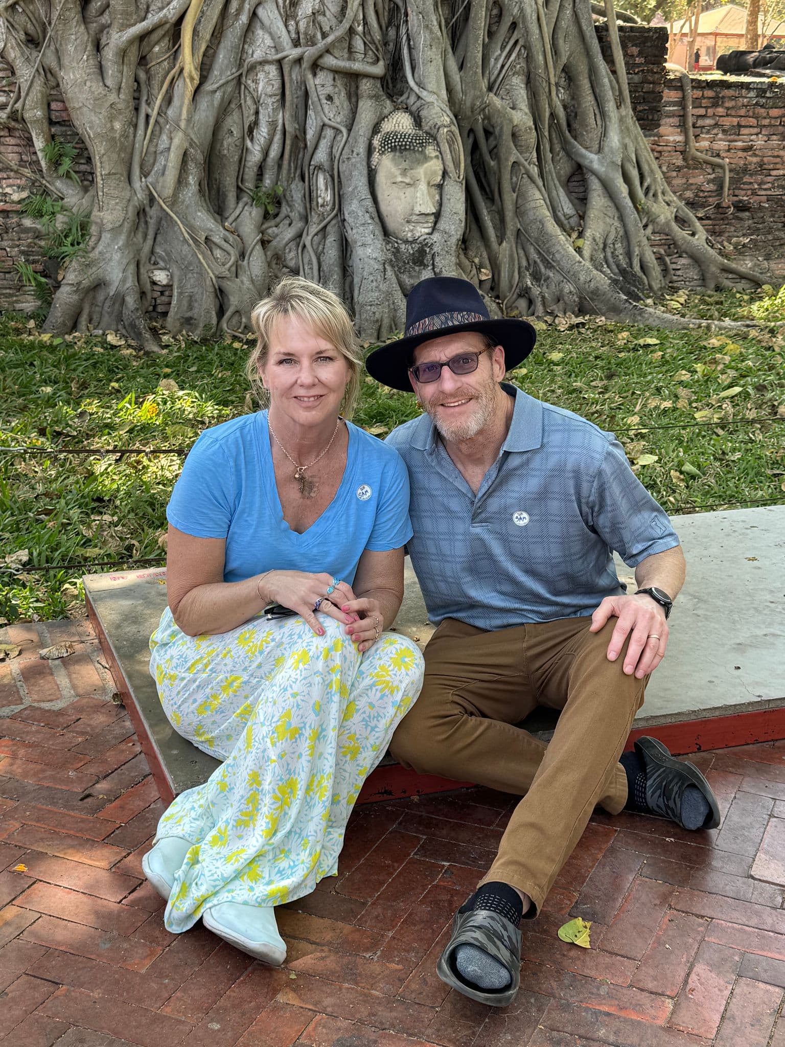 Buddha head entwined in tree roots at Wat Mahathat, Ayutthaya, Thailand, with a couple sitting on a low platform posing for a photo