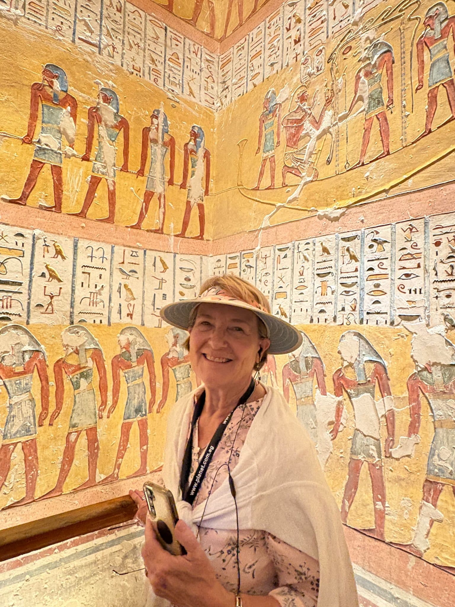 Painted wall reliefs in the Tomb of Ramses IV, Valley of the Kings, Egypt, with a visitor standing and holding a phone during a tour.
