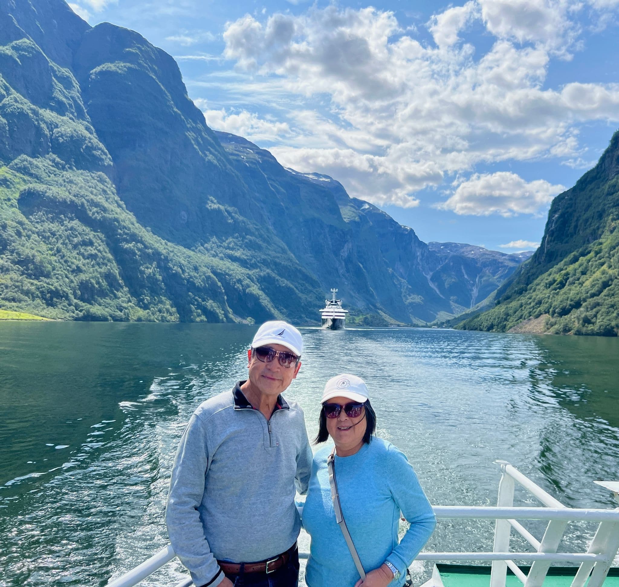 Steep green fjord cliffs and a cruise ship in the water, with two travelers posing on a boat deck in a Norwegian fjord, Norway.