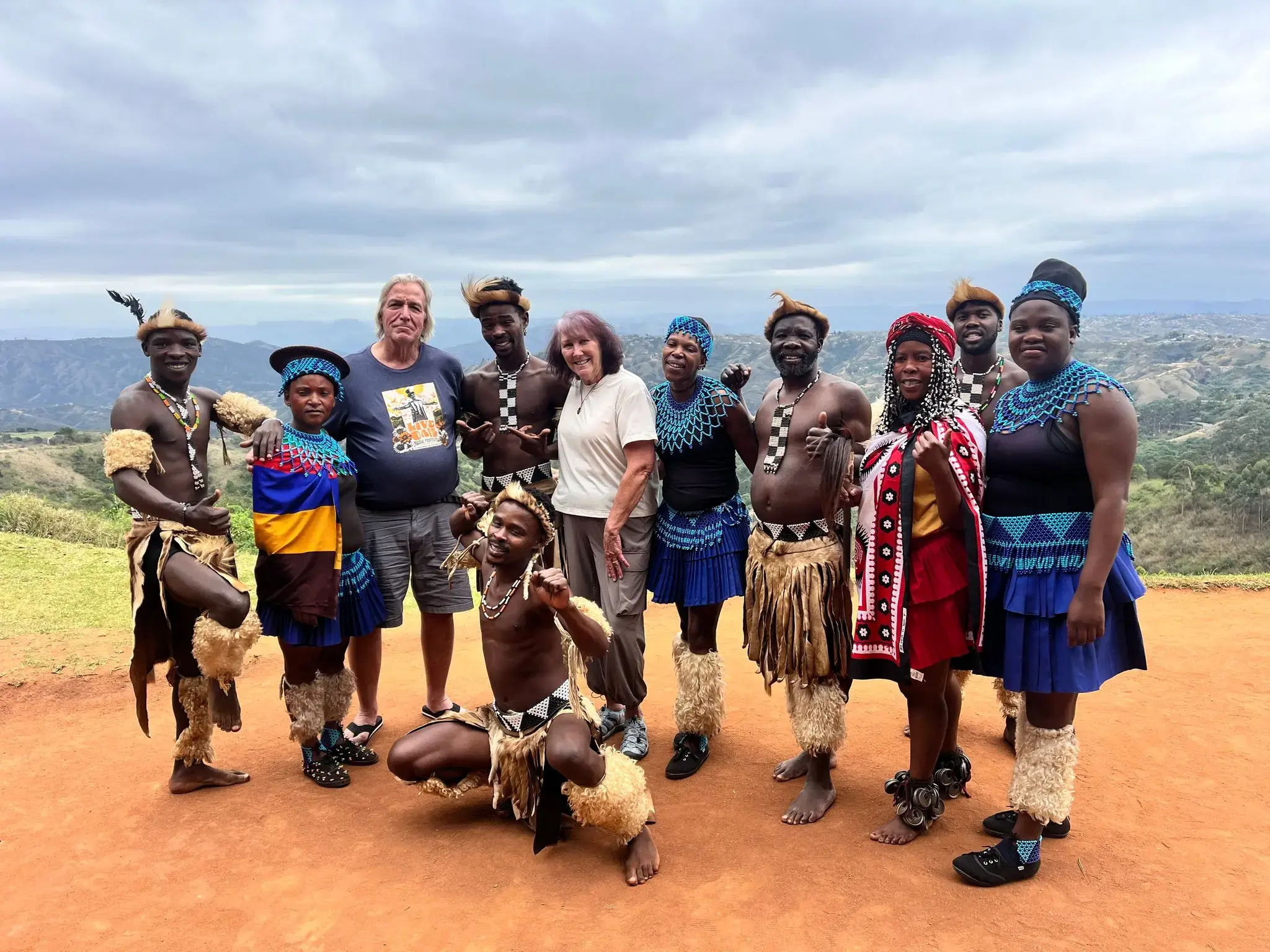 Local dancers and two tourists posing together on a dirt overlook with rolling hills in the background, South Africa.