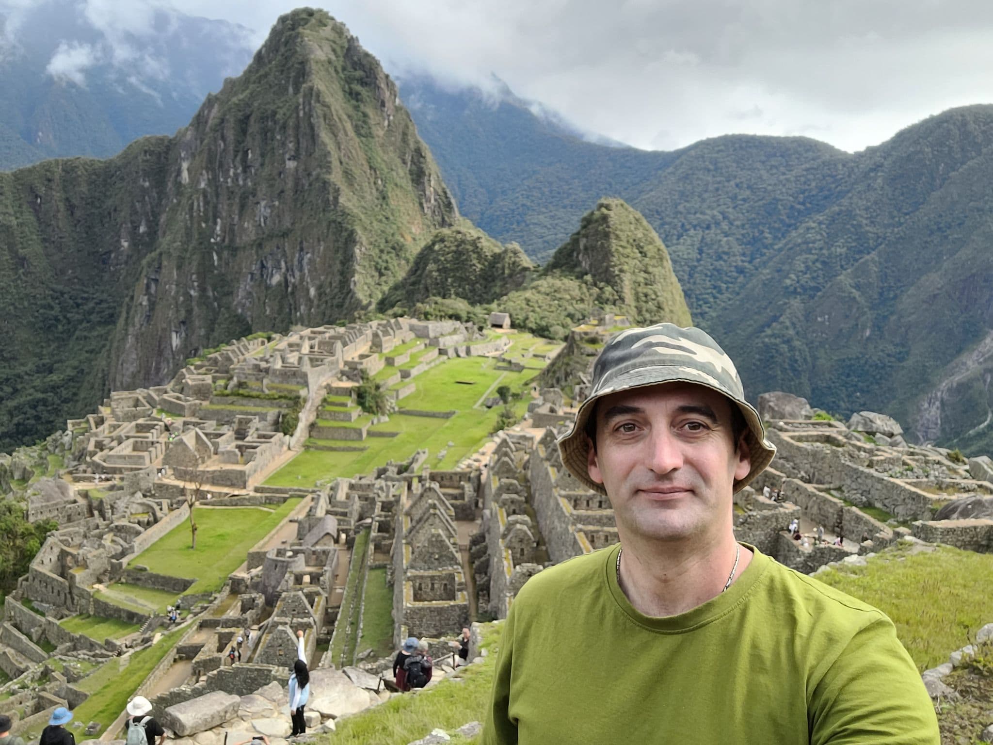 Machu Picchu ruins with a traveler taking a selfie on a grassy terrace, Cusco Region, Peru.
