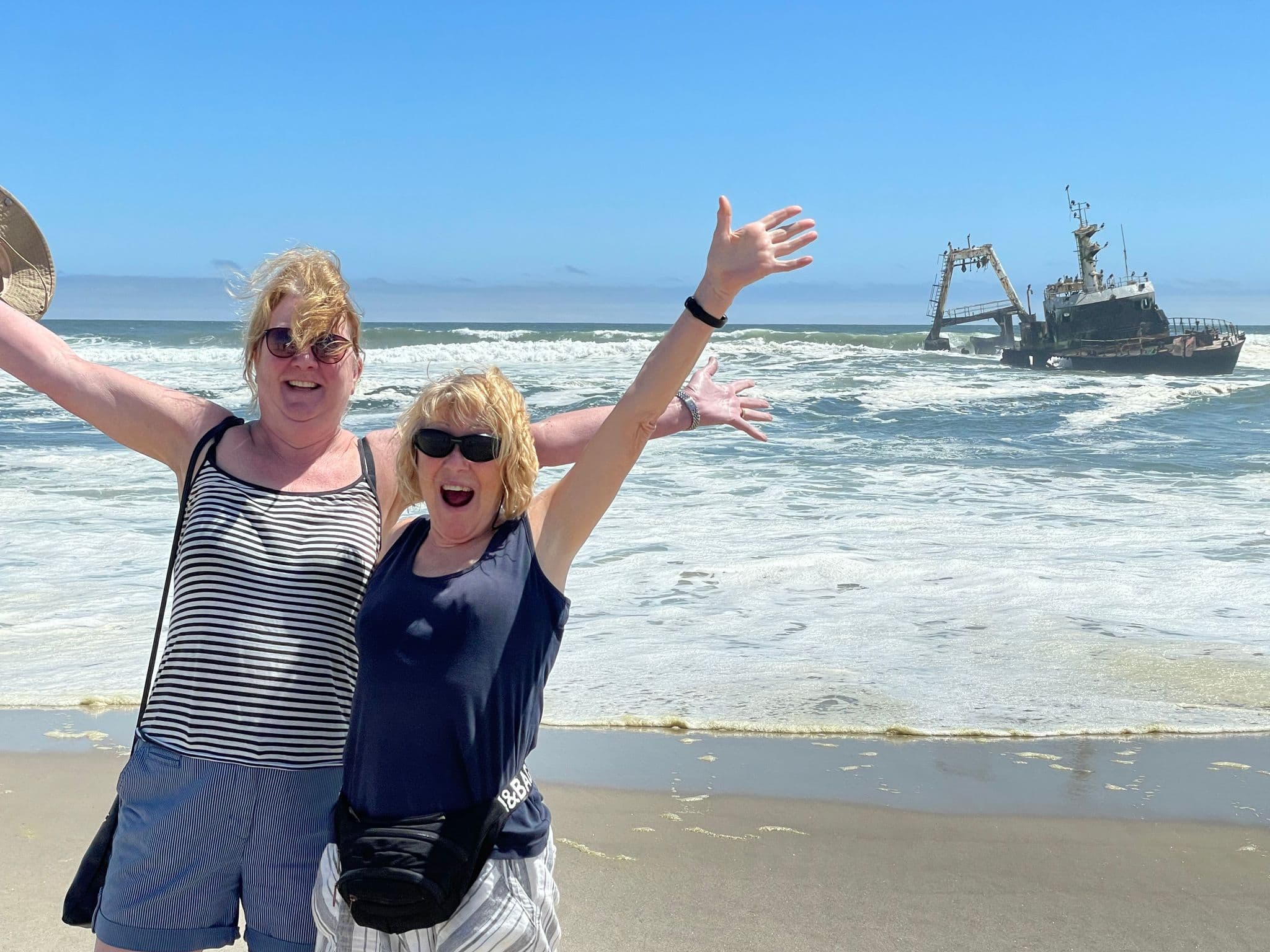 Two women with arms raised on a sandy beach with a rusted shipwreck on the Skeleton Coast at Dorob National Park, Namibia.