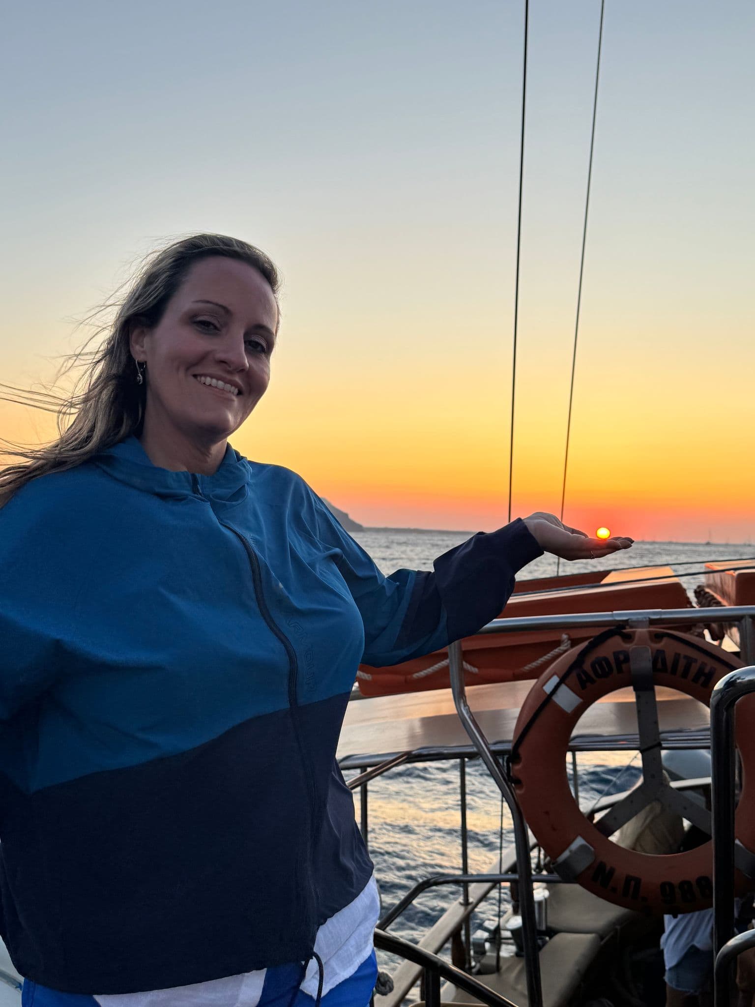 Woman holding the sun in her hand on a sunset dinner cruise at sea, boat railing and life ring visible, Greece.
