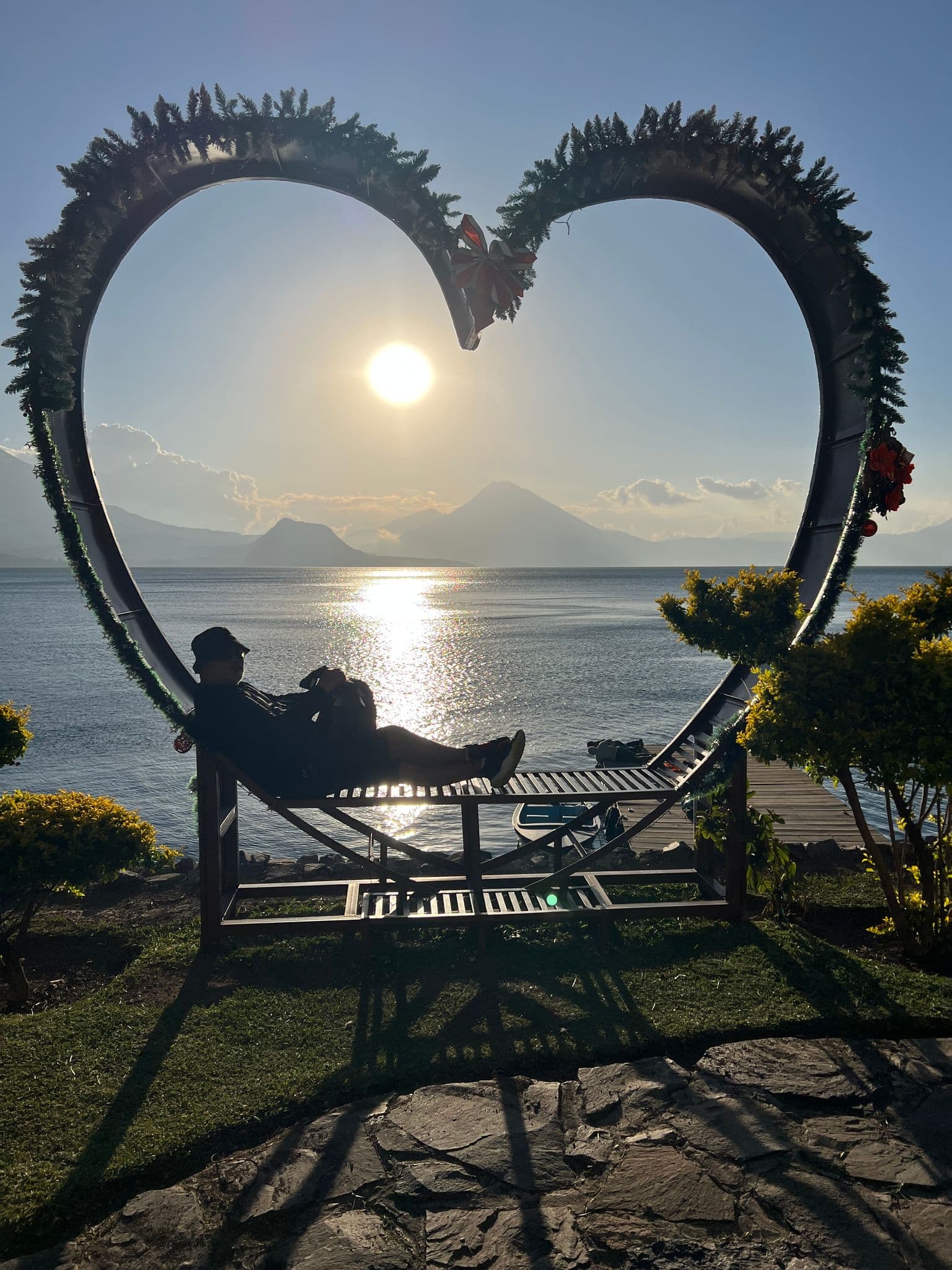 Heart-shaped metal frame framing Lake Atitlán with a reclining person silhouetted against the setting sun and volcano, Guatemala.