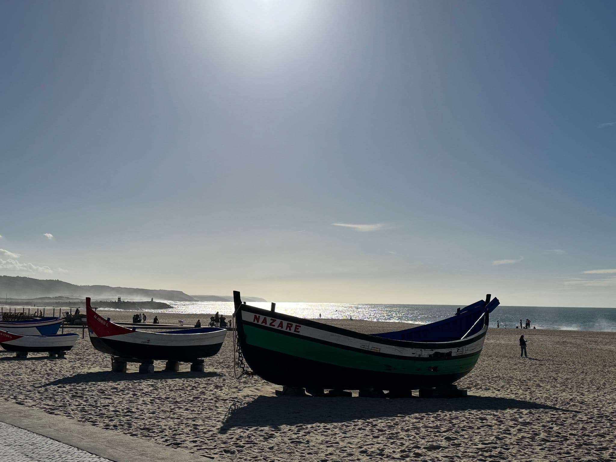 Wooden fishing boats on Nazaré beach, Portugal, with sunlight sparkling on the Atlantic and people walking along the sand.