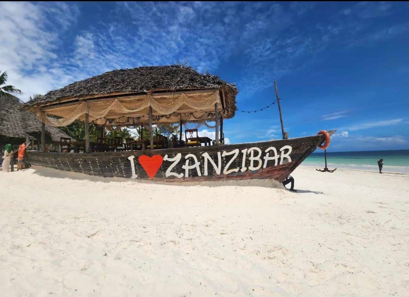 A large wooden boat painted I ? ZANZIBAR on a white-sand beach with a thatched canopy and turquoise ocean, Zanzibar, Tanzania.
