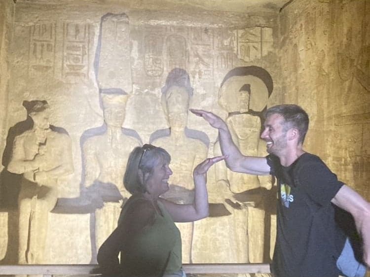 Carved Egyptian temple reliefs behind two people miming a high-five inside a dim temple chamber, Egypt