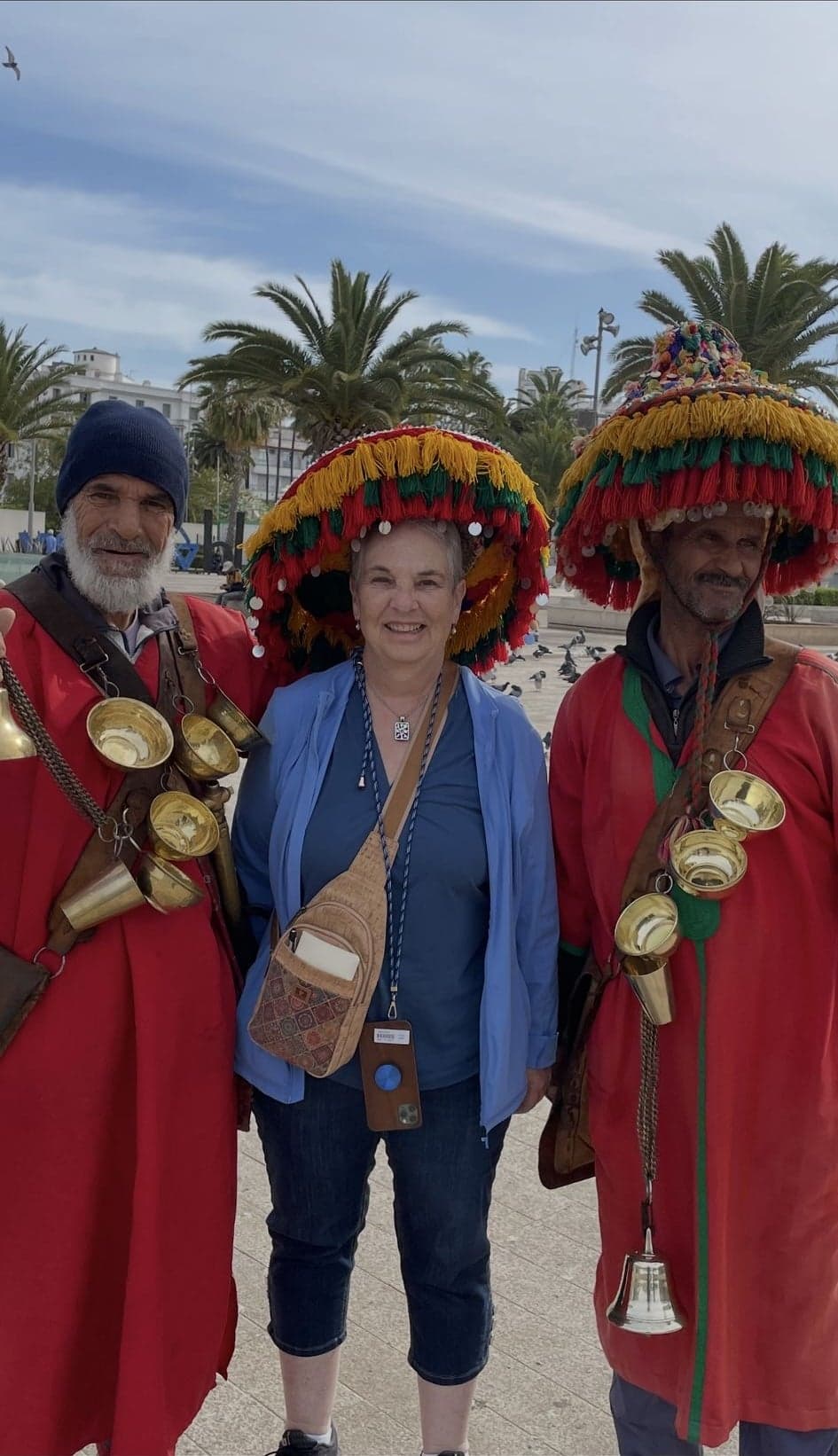 Two traditional Moroccan water sellers in colorful tassel hats with a tourist posing between them in a palm-lined square, Morocco.