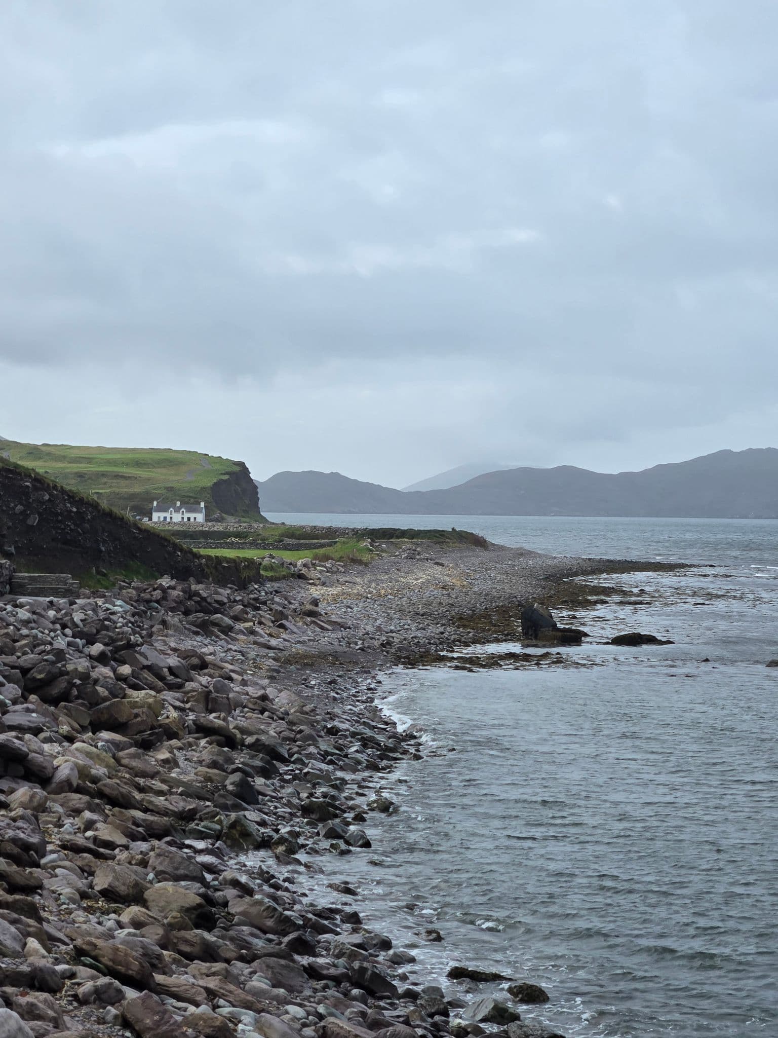 Rocky shoreline with a white cottage on grassy cliffs and distant hills along the coast of Ireland.