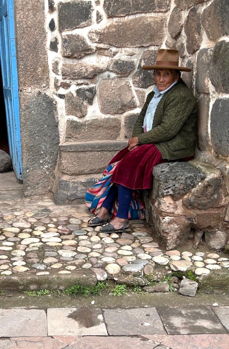 A woman sitting against a stone wall on a cobbled street in a small Andean town, Peru.