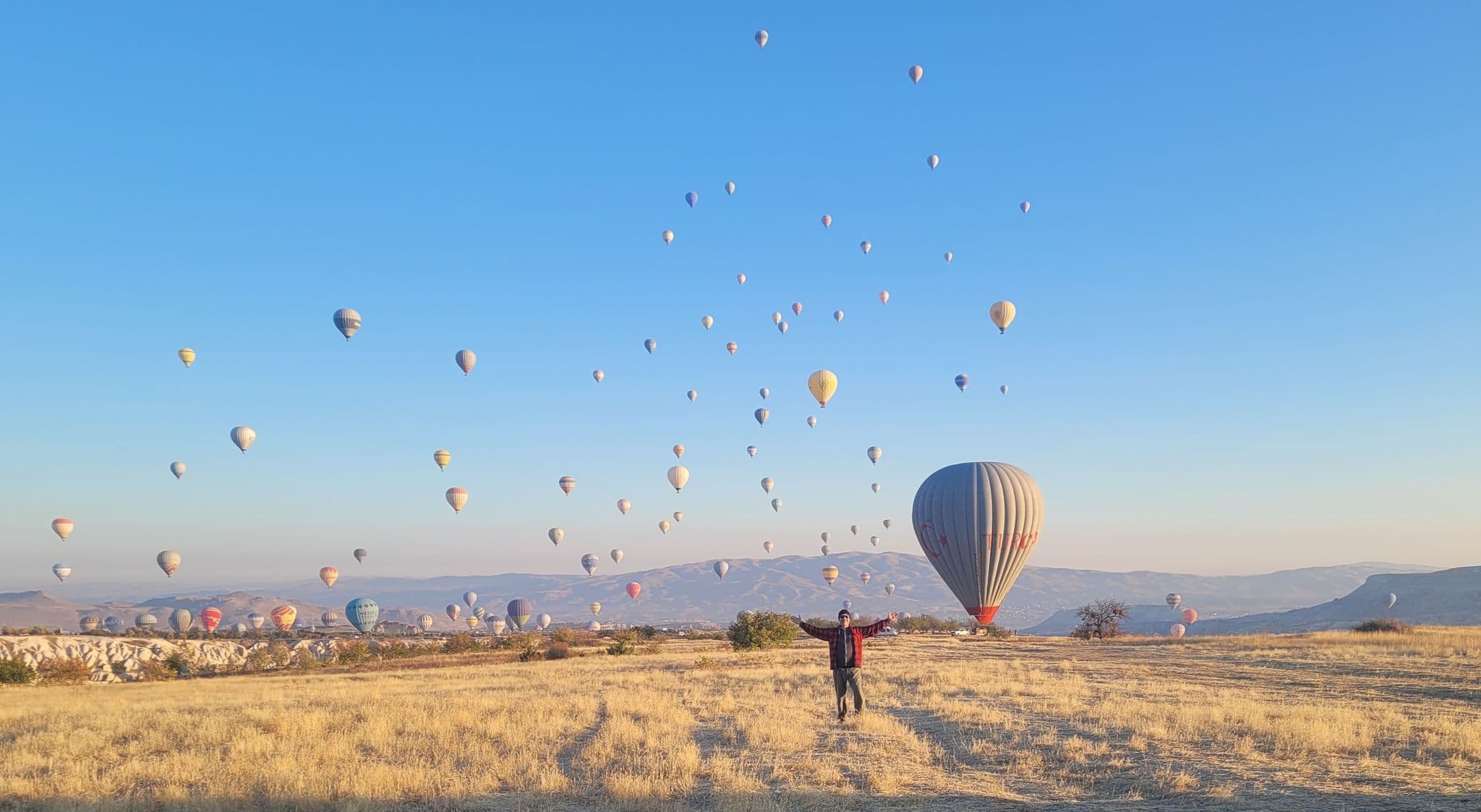 Hot air balloons filling the sky over Cappadocia (Göreme), Turkey, while a person stands in a dry field with arms outstretched.