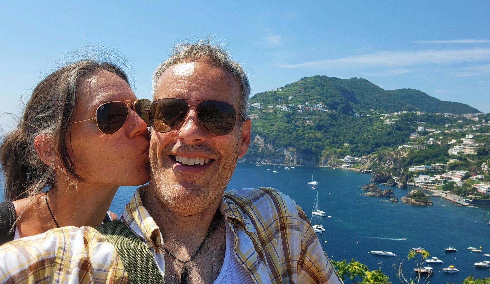 View from Aragonese Castle in Ischia, Italy, of a man smiling while his partner kisses his cheek with the bay and boats below.