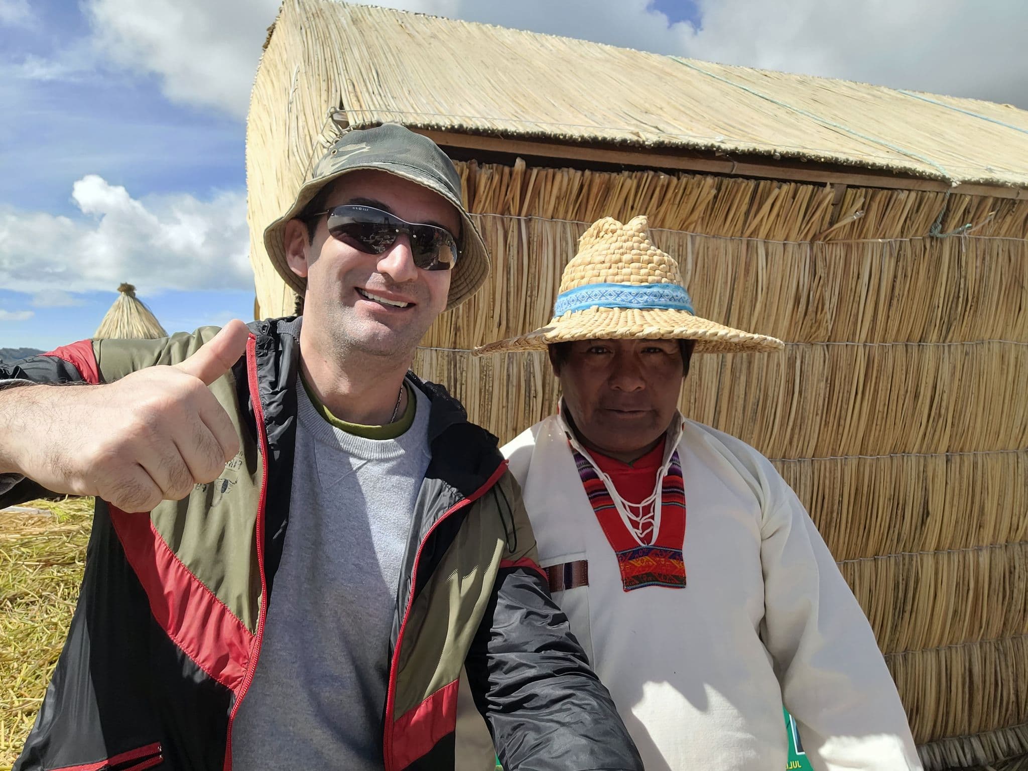 Reed hut on the Uros Floating Islands with a tourist giving a thumbs-up and a local man in a straw hat posing for a selfie, Lake Titicaca, Peru.