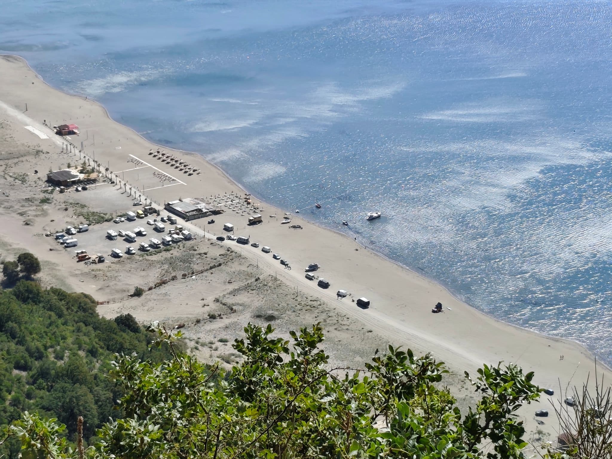Sandy coastline with parked cars, beach umbrellas, and a small boat seen from a hillside viewpoint.