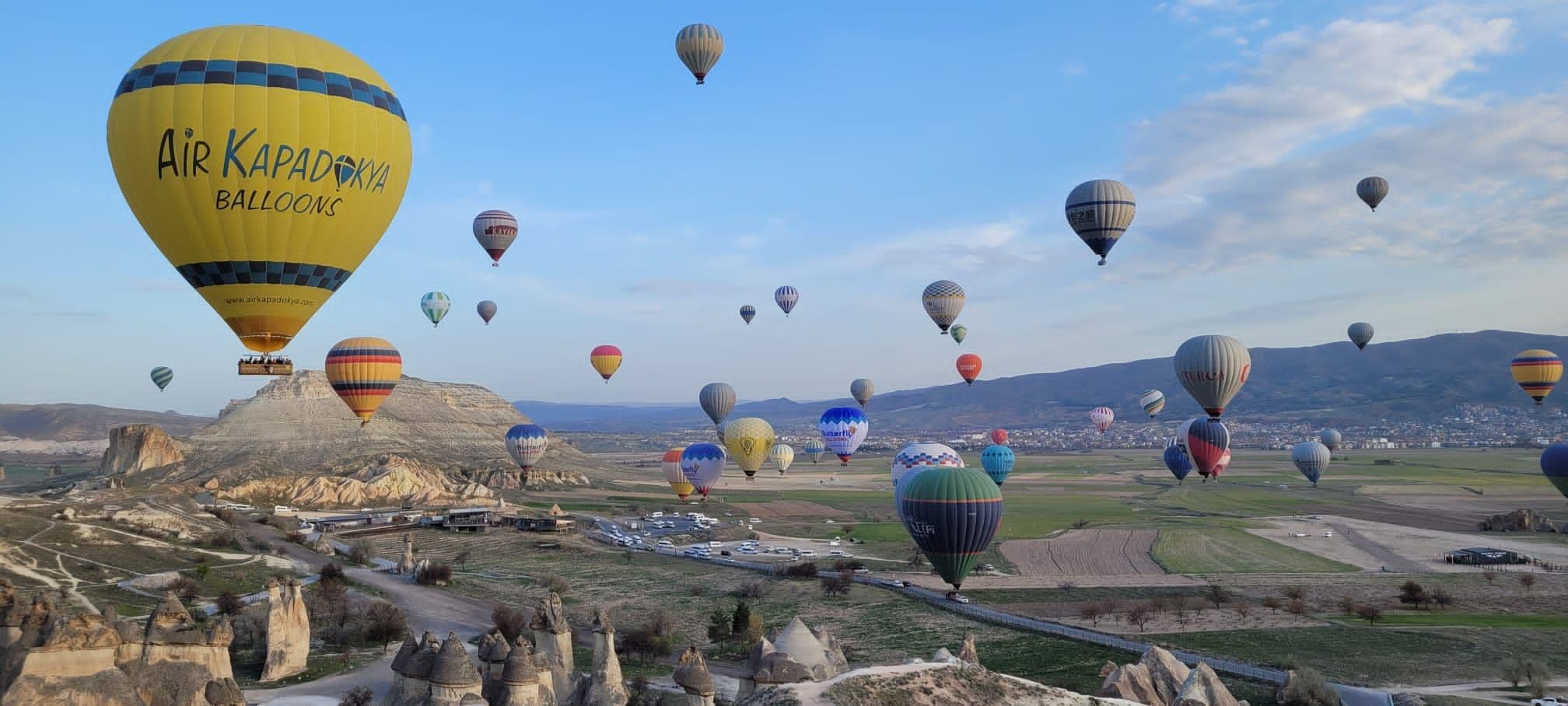 A large yellow Air Kapadokya hot-air balloon above Cappadocia's fairy chimneys with dozens of colorful balloons over the valleys, Turkey.