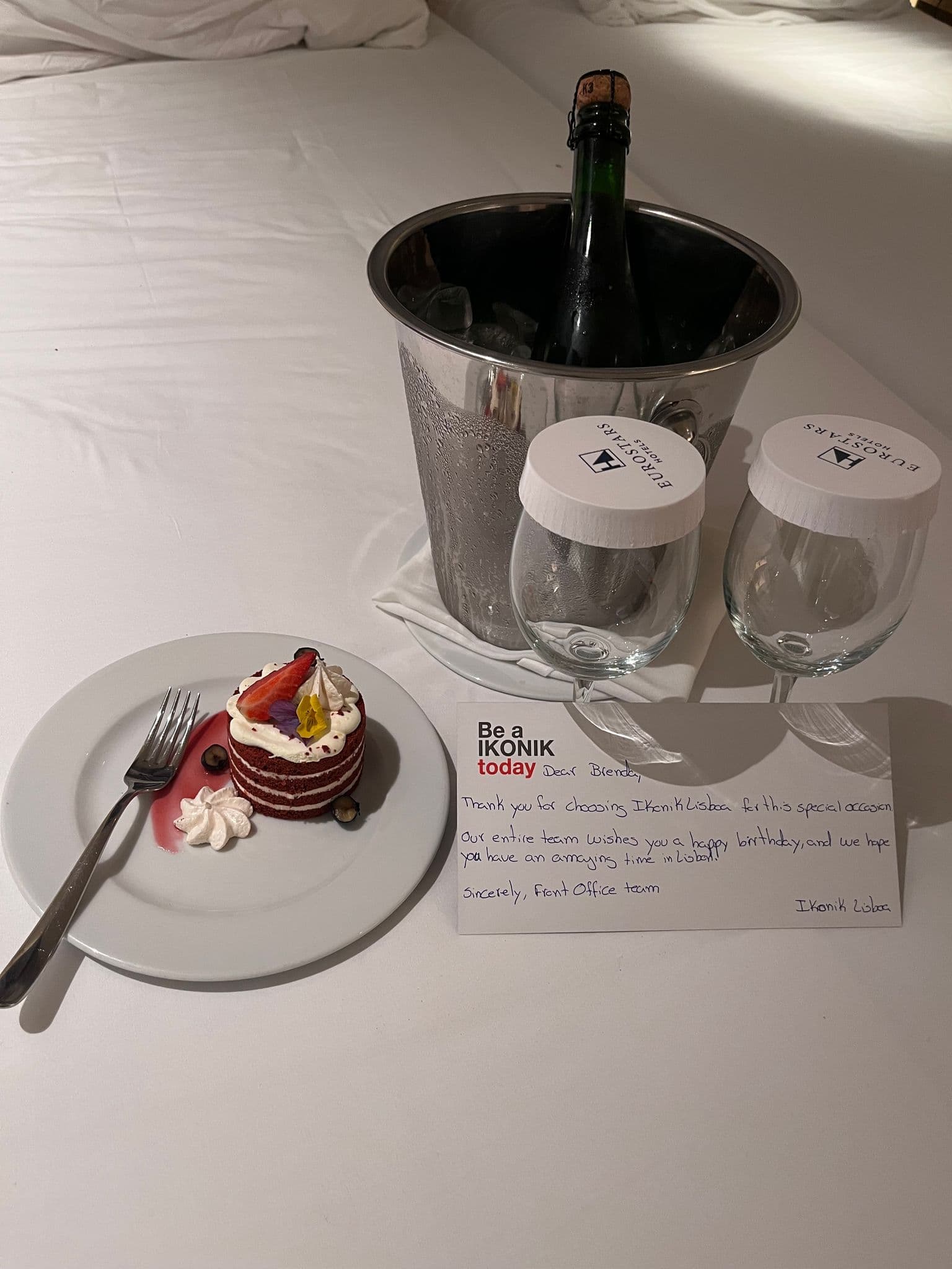 Champagne bottle chilling in an ice bucket with two covered wine glasses, a small red-velvet cake, and a handwritten hotel birthday note on a bed in Lisbon, Portugal.
