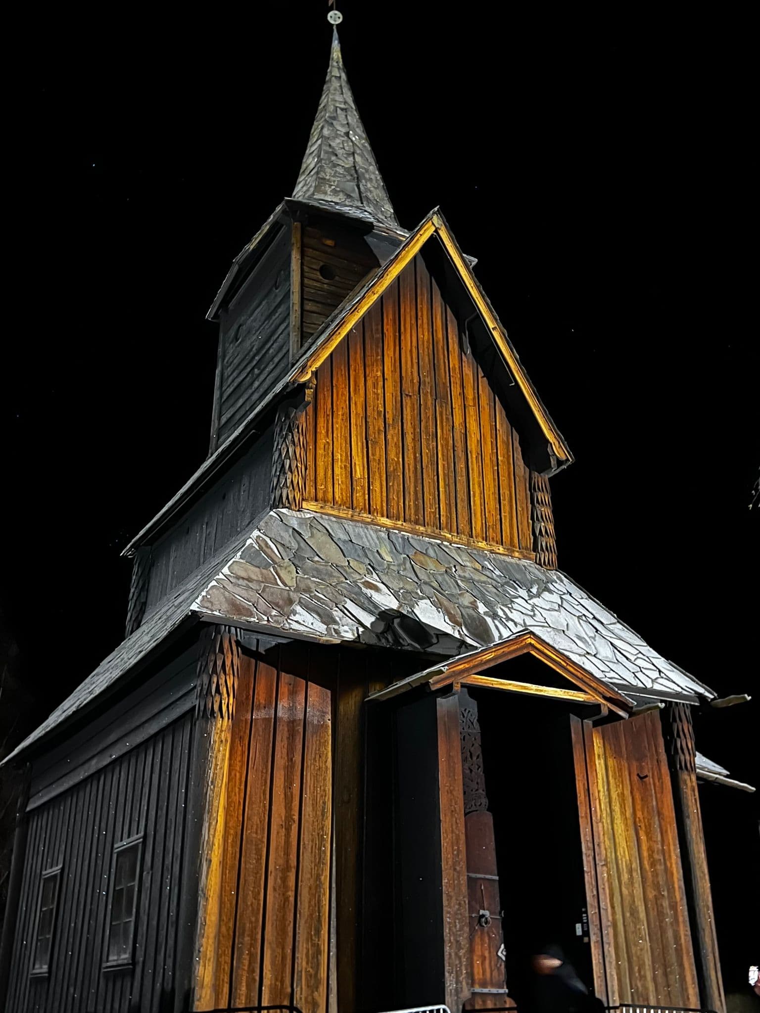 Wooden stave church illuminated at night against a black sky, showing a carved entrance and steep spire, Norway.