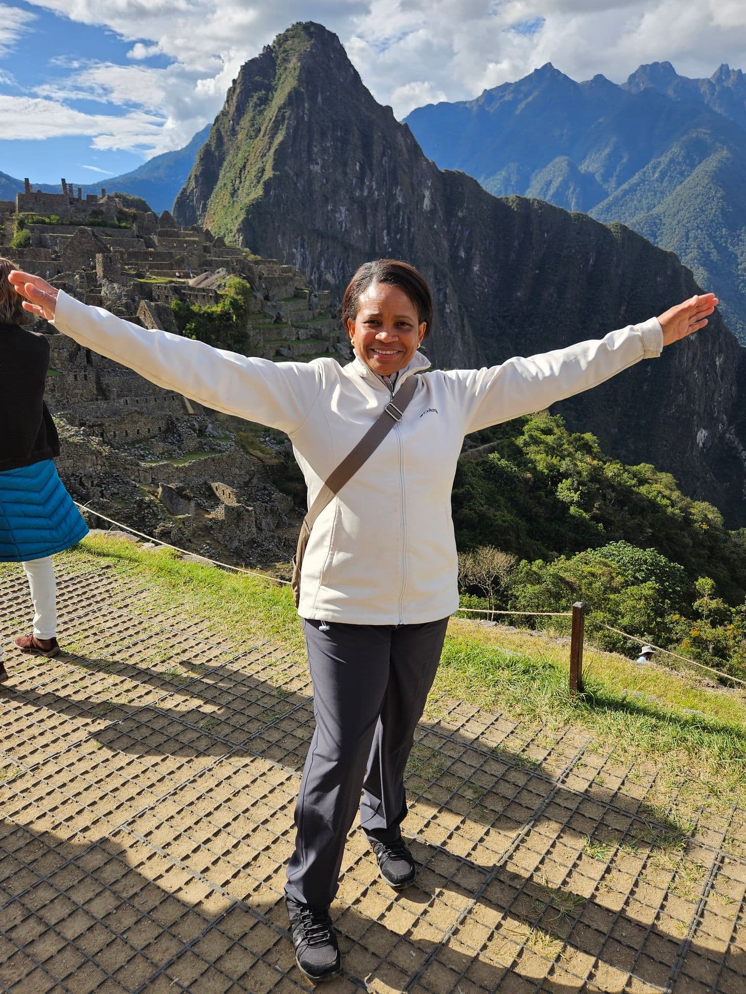 Machu Picchu ruins with Huayna Picchu in the background and a woman standing with arms outstretched on a viewing terrace, Cusco Region, Peru.