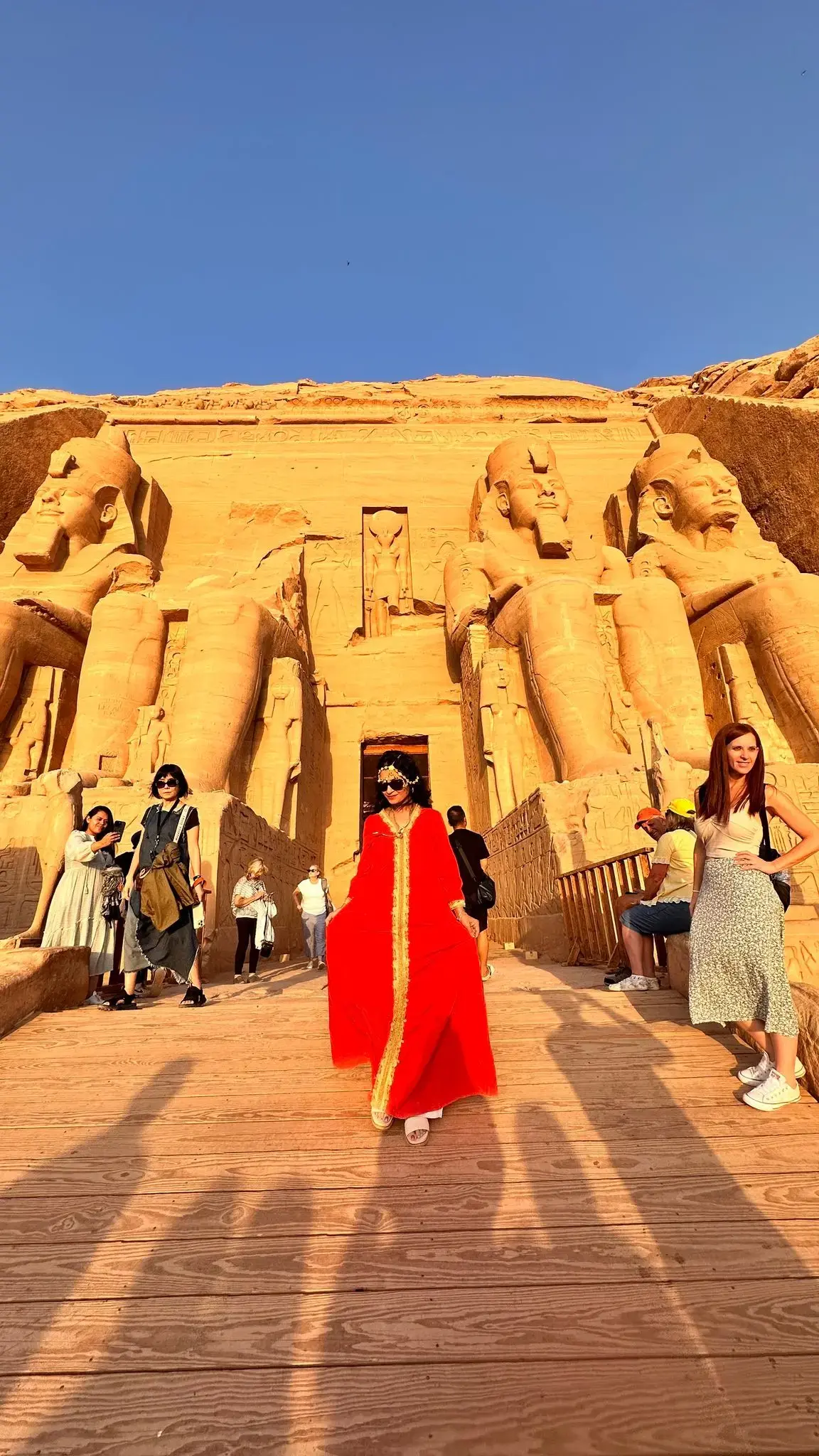 Abu Simbel temple facade with giant seated statues and people, including a woman in a red dress on a wooden ramp, Egypt.