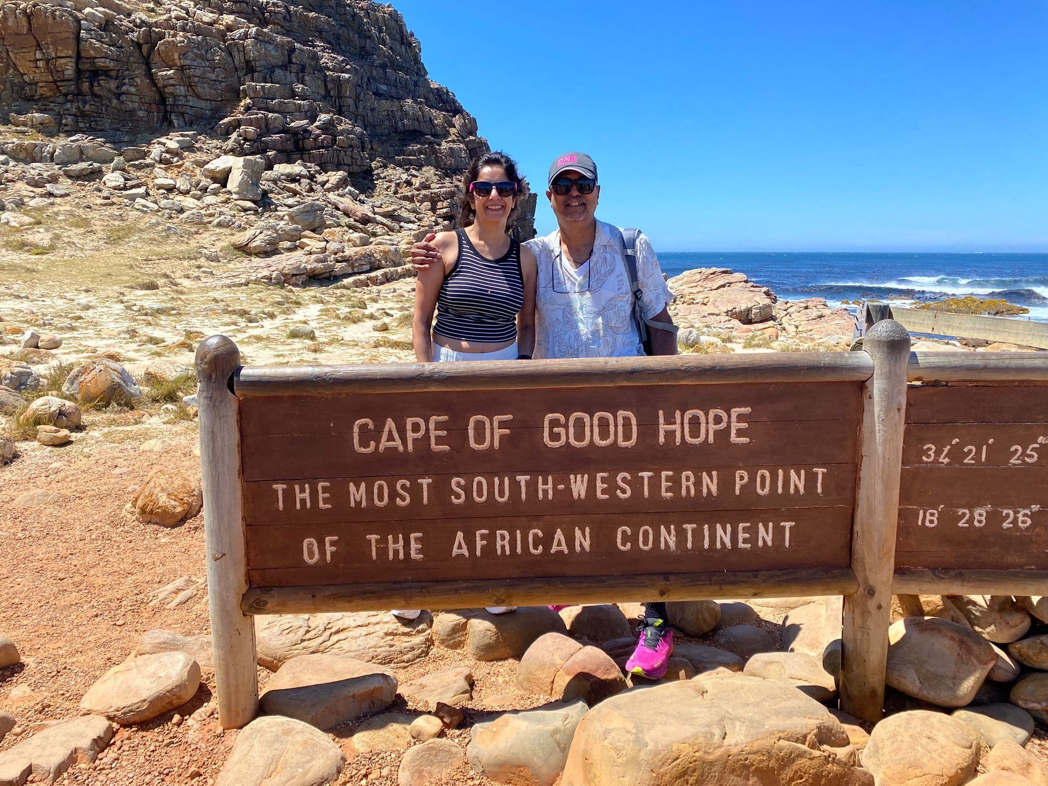 Cape of Good Hope sign with two travelers posing behind it on the rocky Atlantic coast, Western Cape, South Africa.