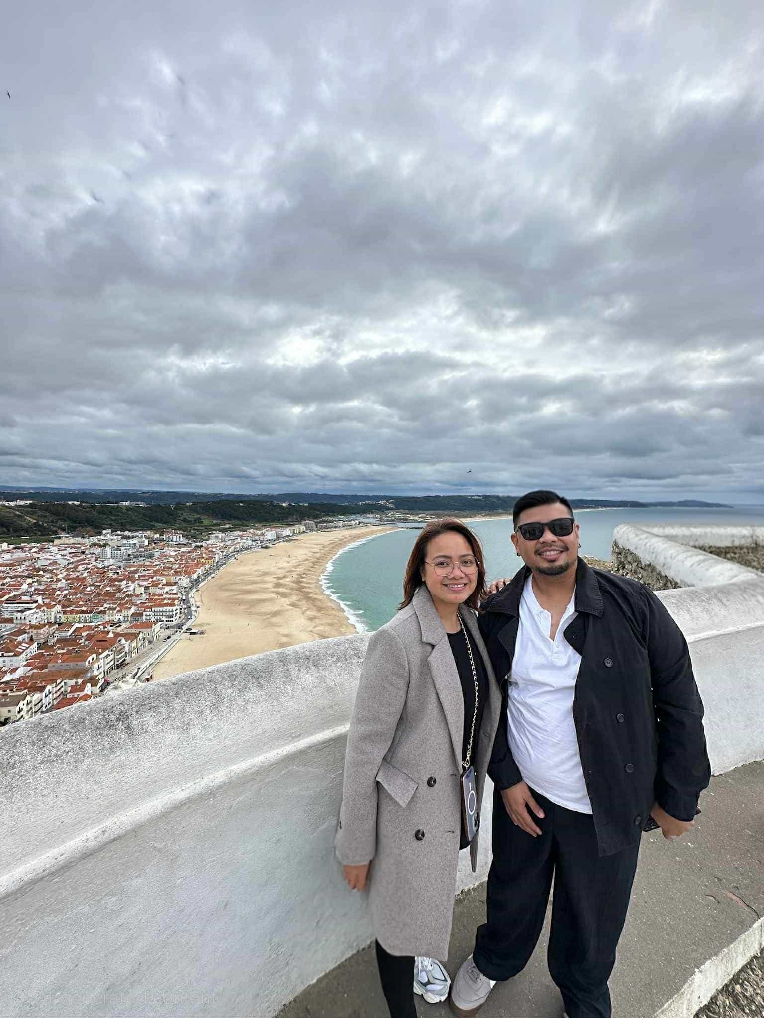 Praia de Nazaré beach and town seen from a cliffside viewpoint in Nazaré, Portugal, with two travelers posing at a white wall.