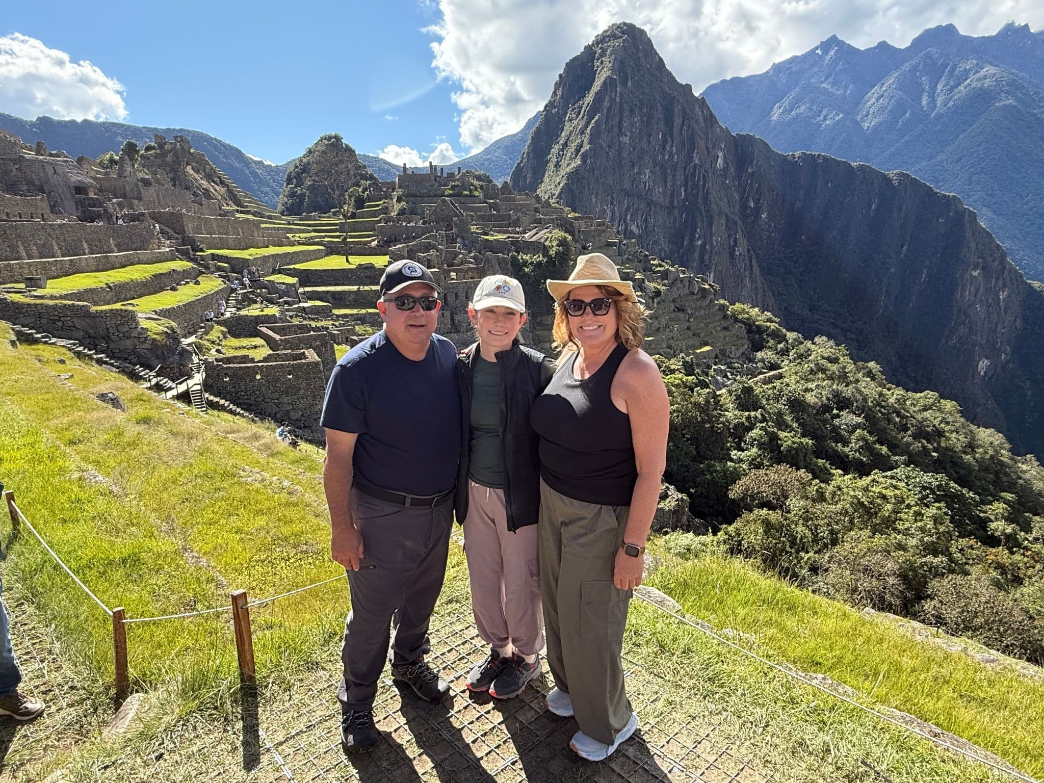 Machu Picchu ruins with three family members standing together on a grassy terrace, Huayna Picchu and the Andes behind, Peru.
