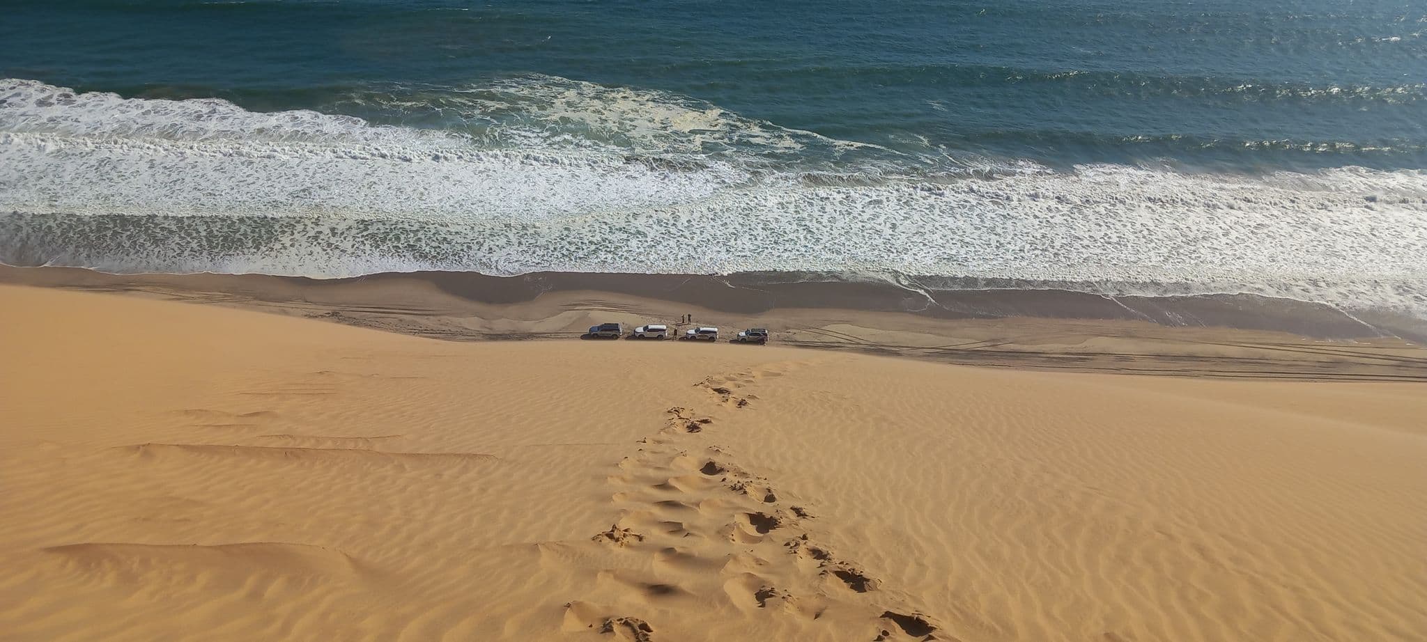 Steep sand dune with footprints descending to the Atlantic shoreline where several vehicles are parked on a Namib Desert beach, Namibia.