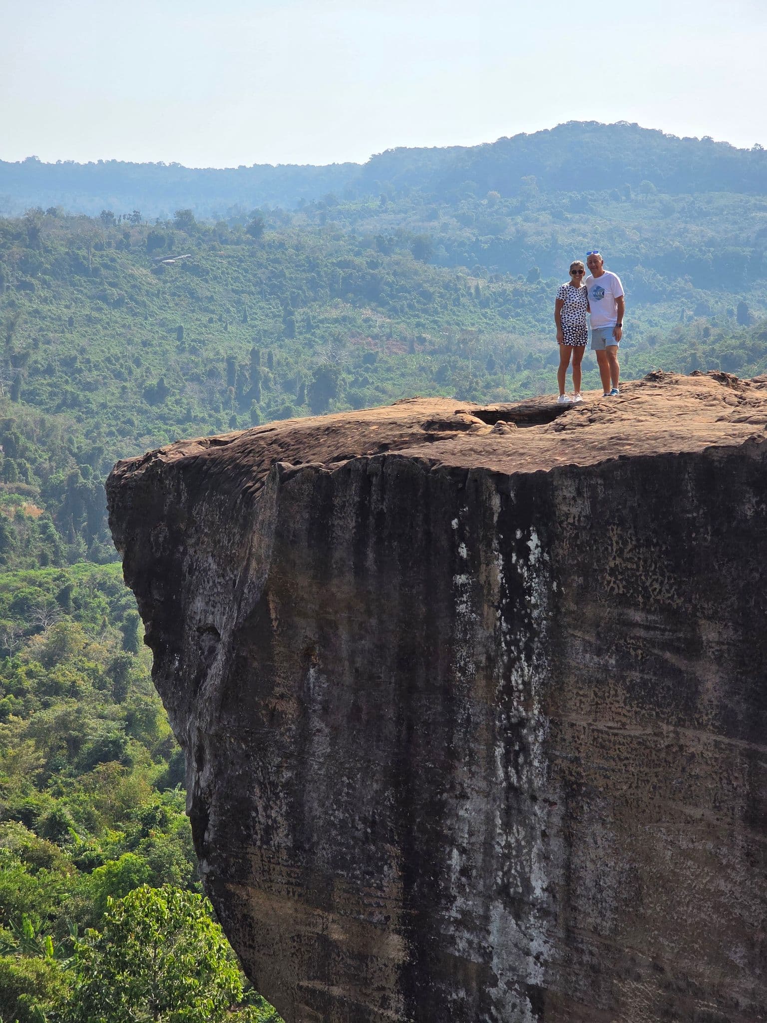 Large cliff with two people standing near the edge, overlooking a green forested valley and distant hills.