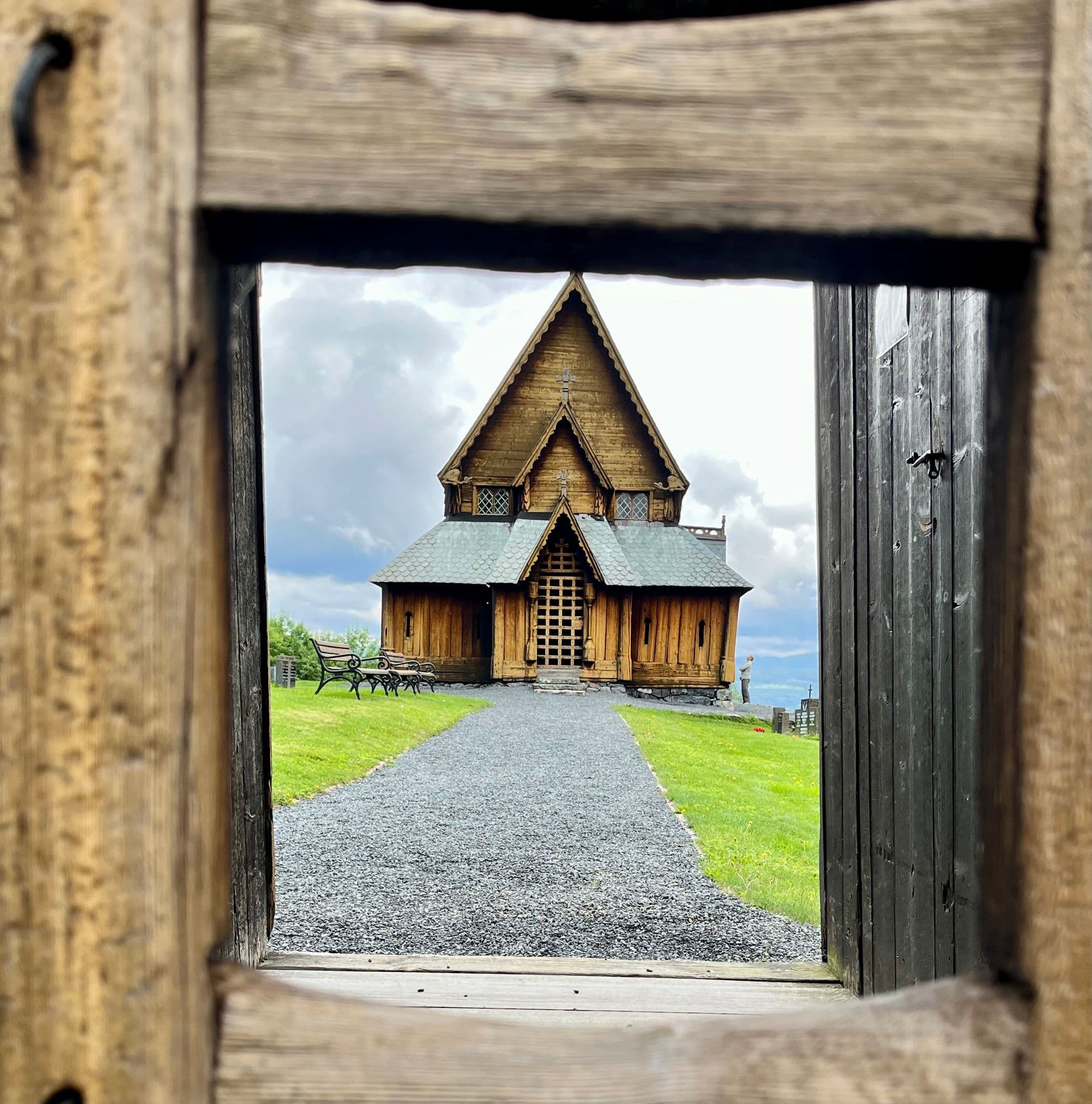 Wooden stave church framed through a gate, standing at the end of a gravel path on a grassy hill, Norway.