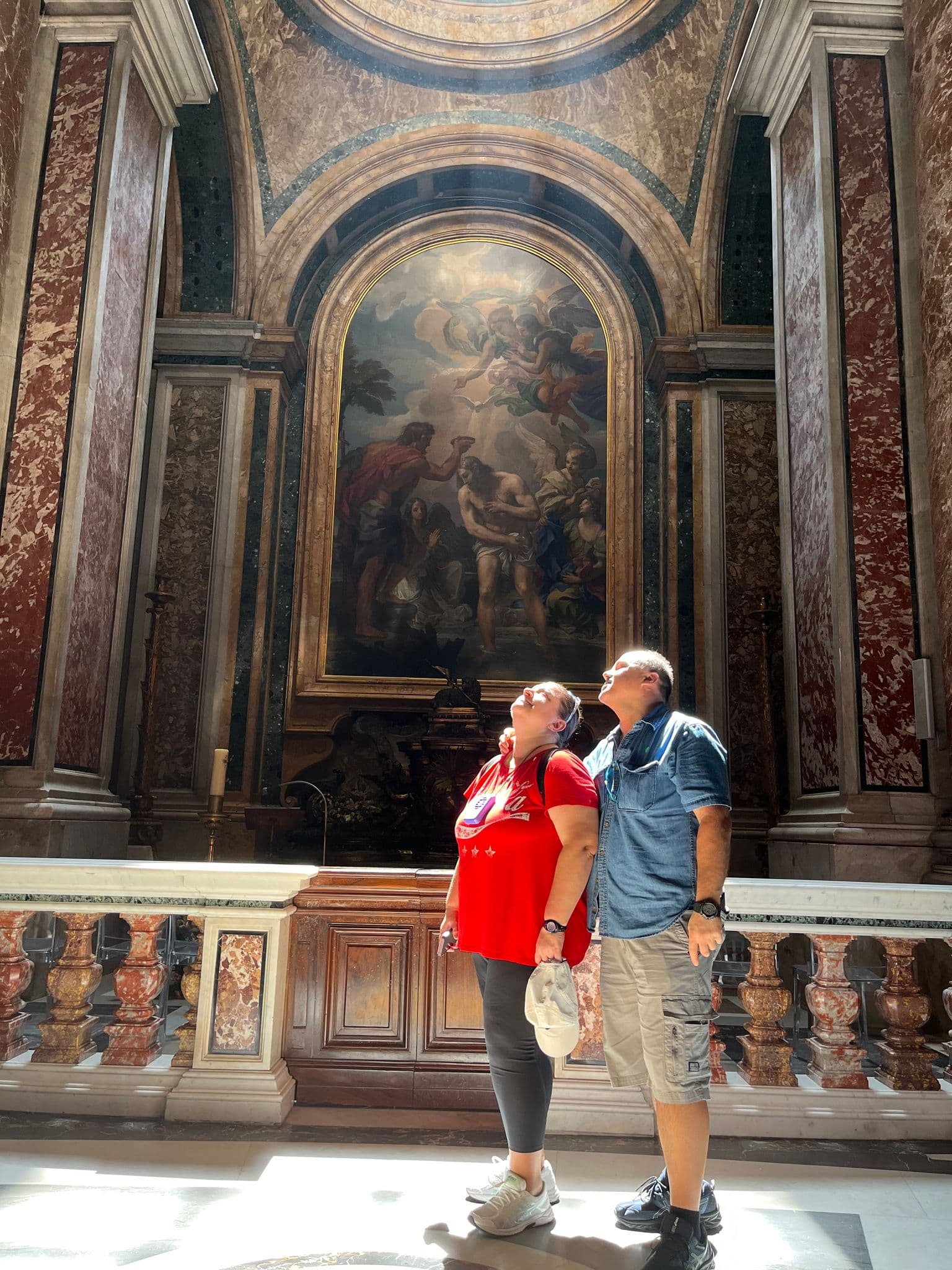 Two travelers looking up at a large baroque altar painting in a sunlit church interior, Italy