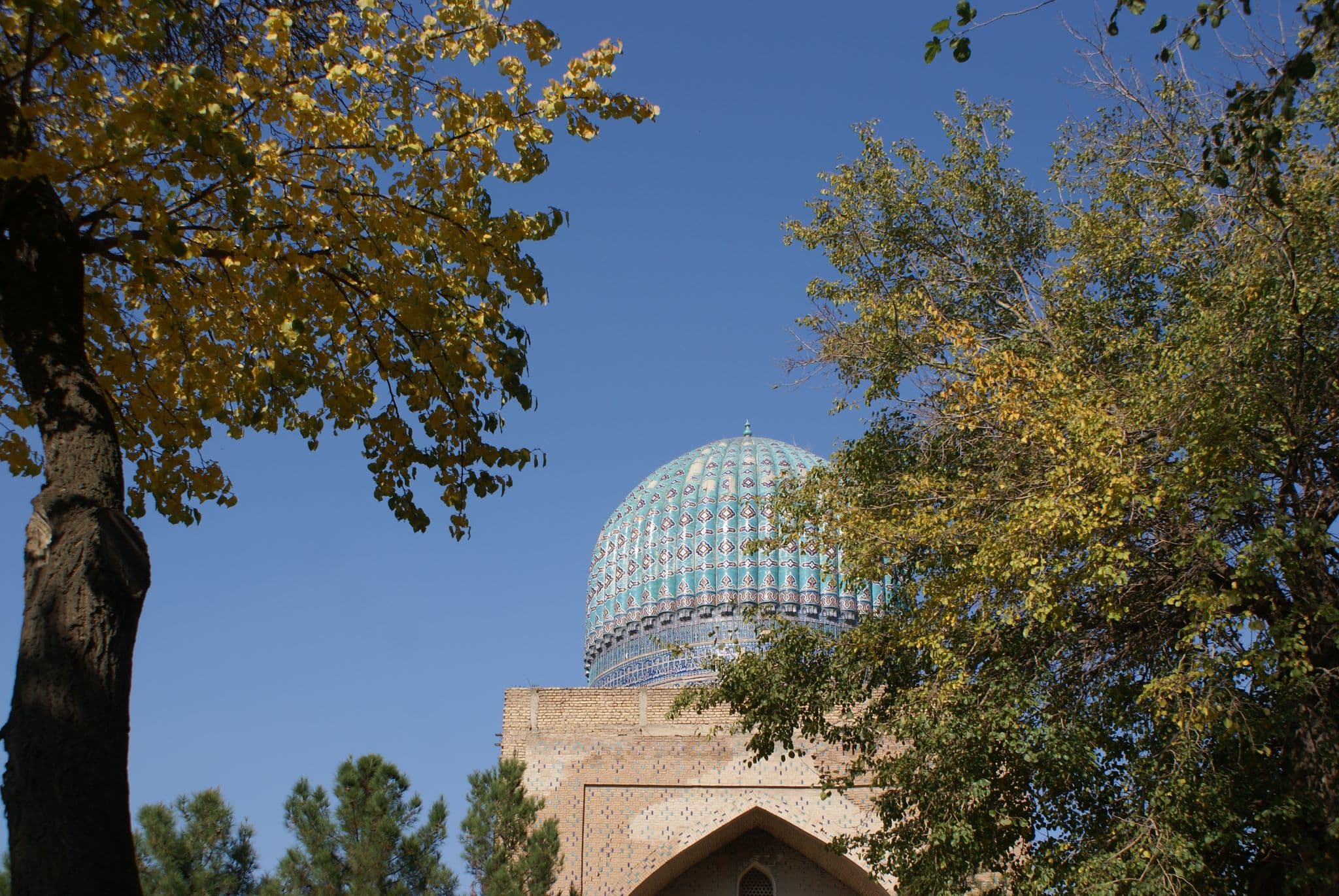 Turquoise tiled dome of Gur-e-Amir Mausoleum rising above a brick portal, framed by trees in Samarkand, Uzbekistan.