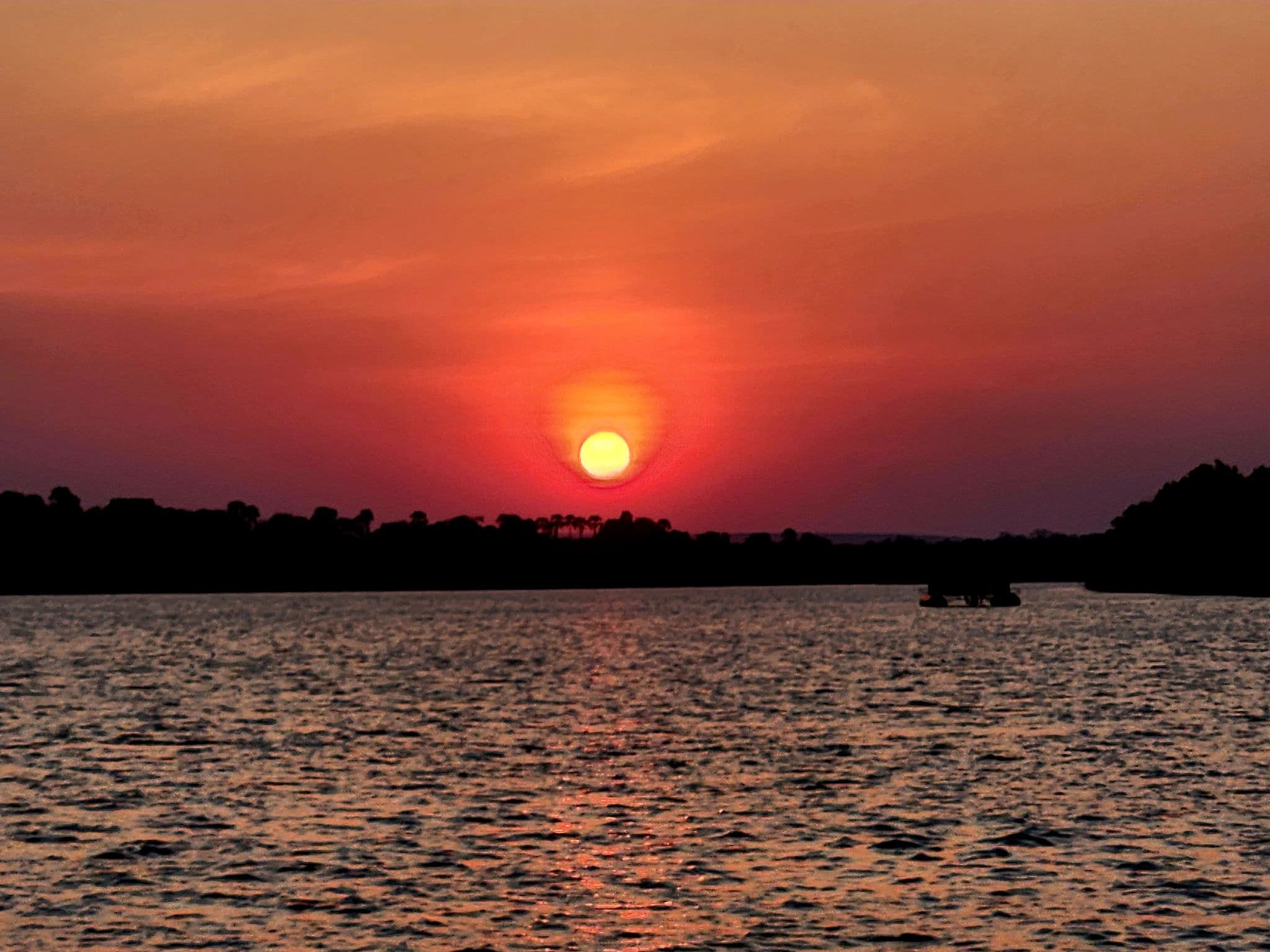 Sun setting over a river with a silhouetted boat on a river cruise, trees on the horizon, South Africa.