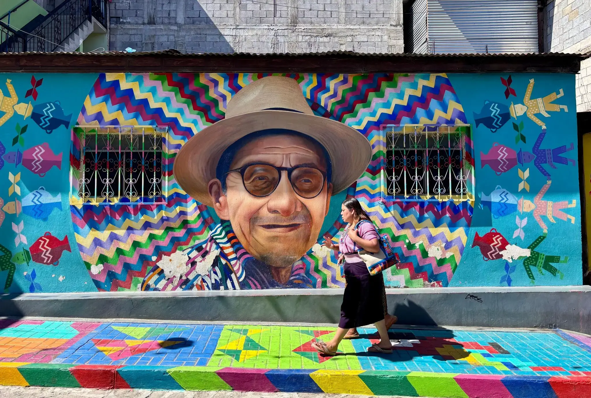 Large painted mural of a man wearing a hat on a colorful wall, with a woman walking on the painted sidewalk in San Juan La Laguna, Guatemala.
