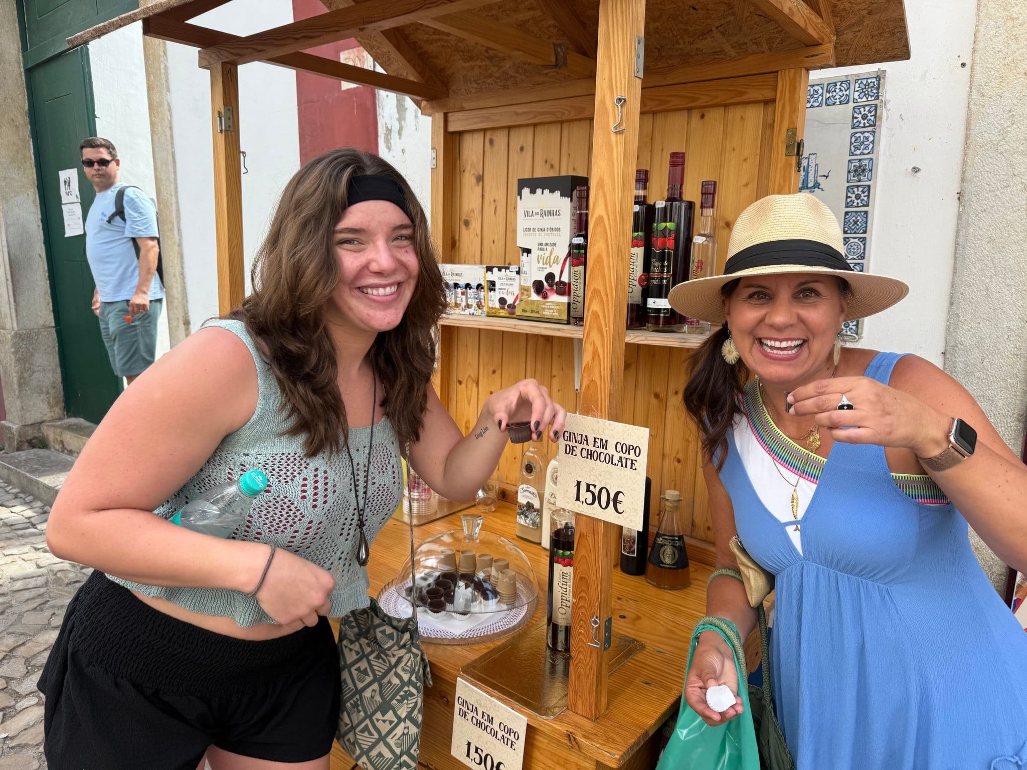 Two women sampling ginja cherry liqueur served in chocolate cups at a wooden market stall in Óbidos, Portugal.