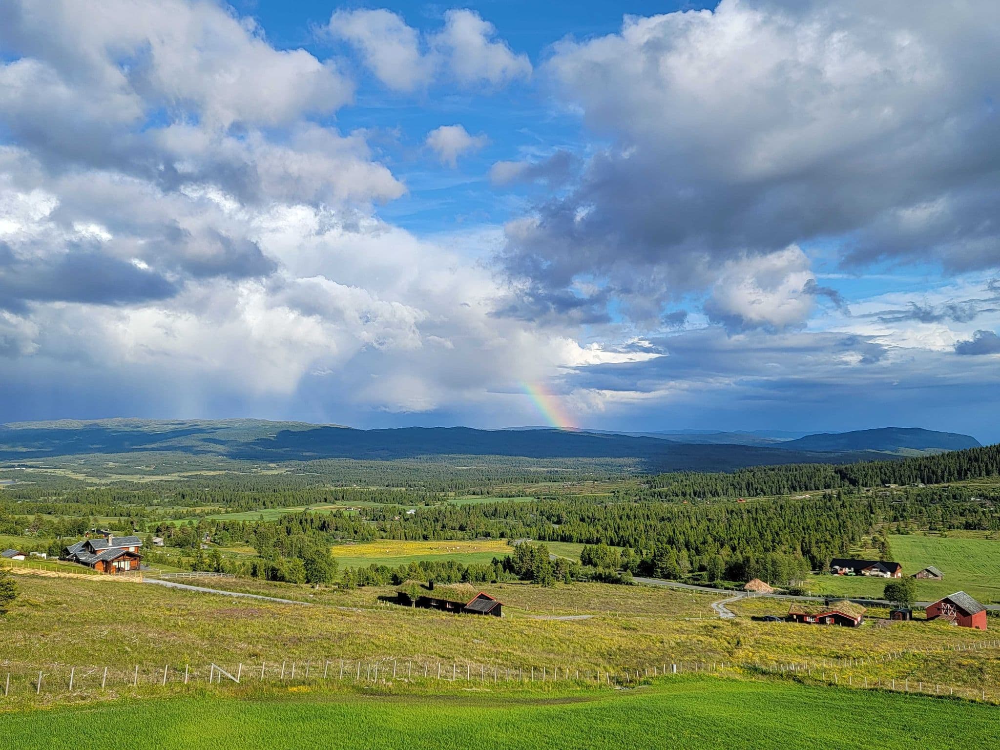 Rolling green fields and scattered red cabins under a cloudy sky with a rainbow near Oslo, Norway.