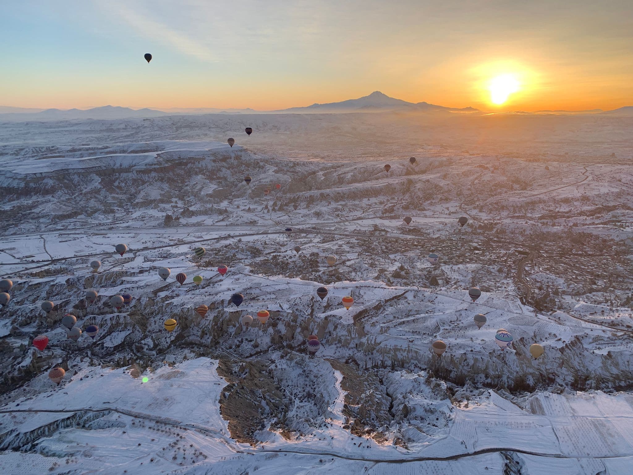 Hot air balloons floating over the snow-covered valleys of Cappadocia at sunrise with a distant mountain in Turkey