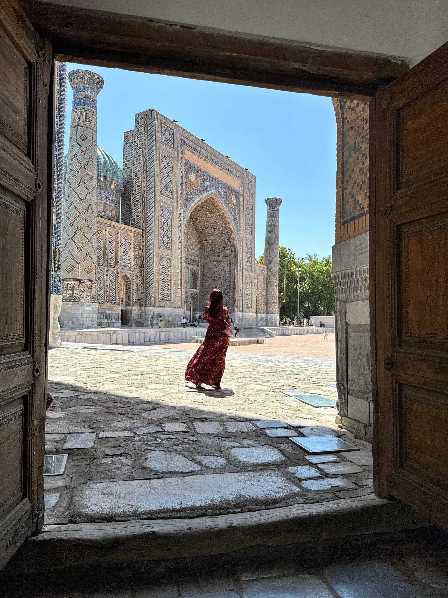 Registan's ornate madrasa framed by a wooden doorway as a person in a red dress walks the courtyard, Samarkand, Uzbekistan.
