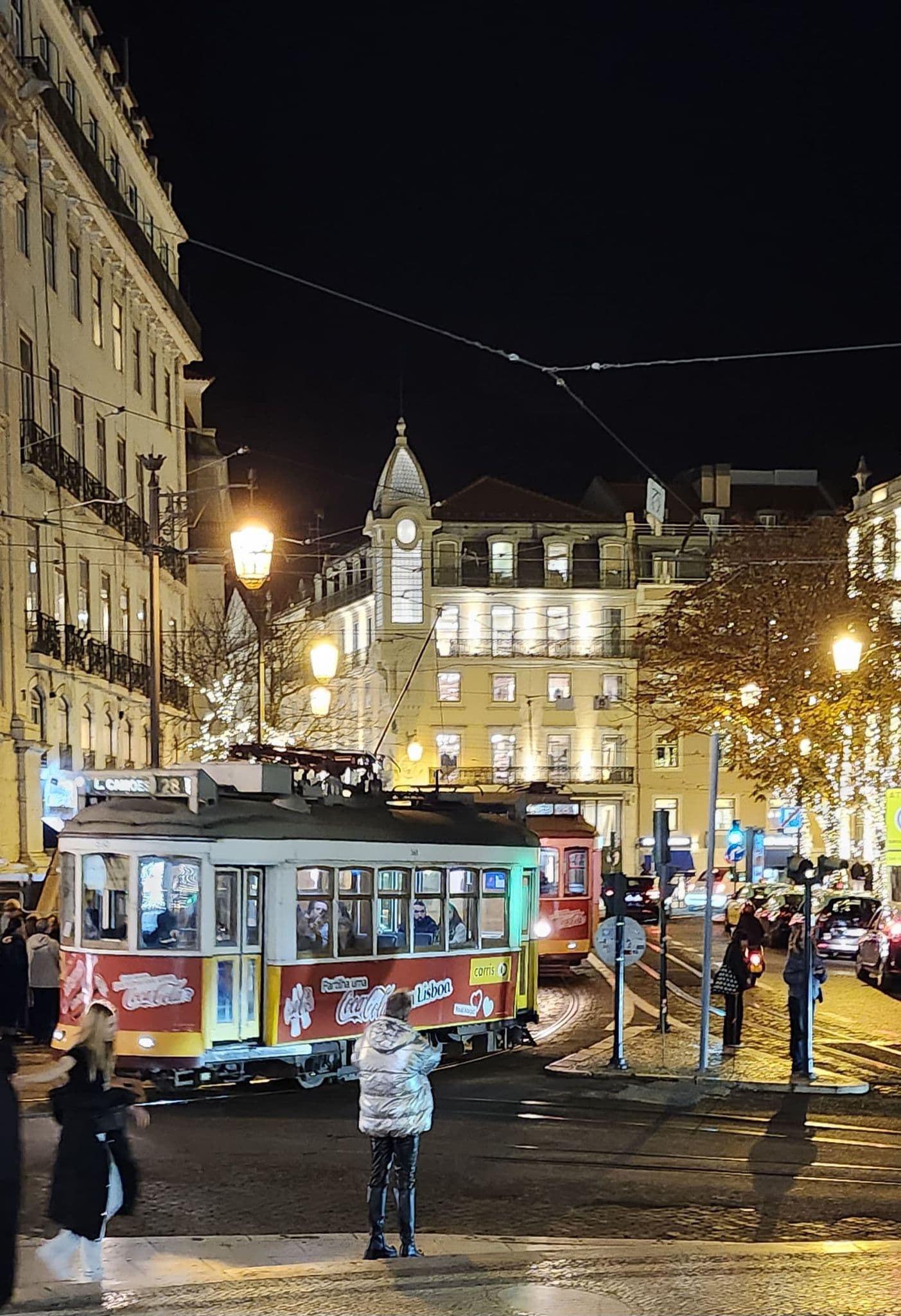 Historic Lisbon tram (Tram 28) passing a lit city street at night in Lisbon, Portugal, with people nearby.