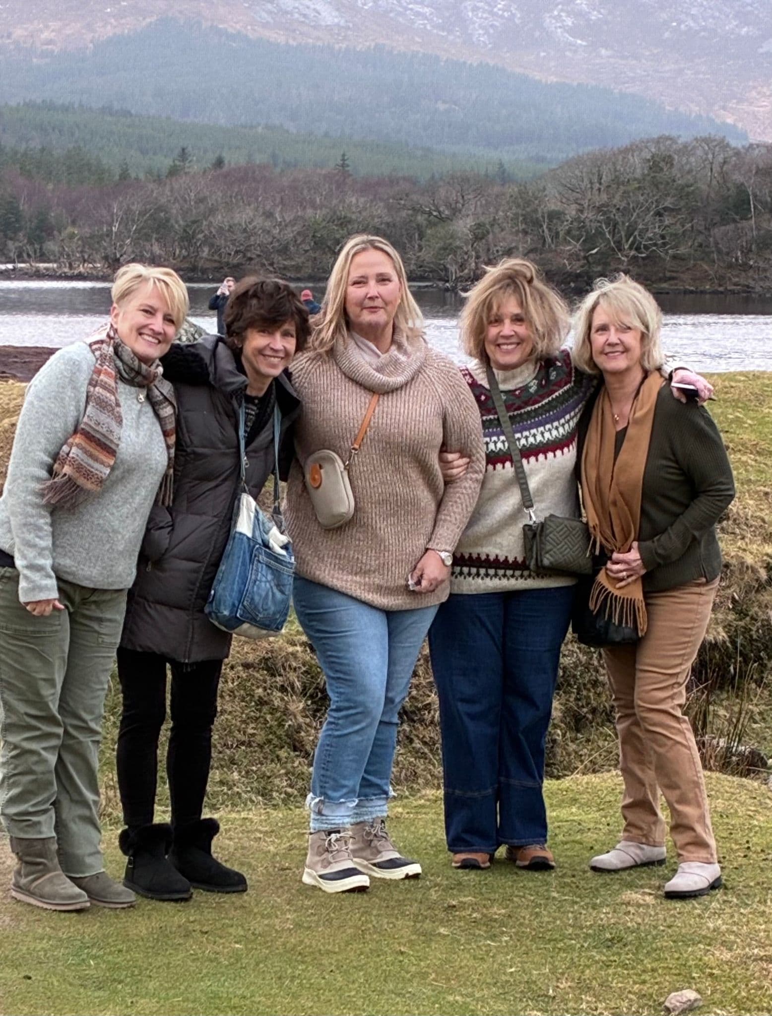 Five women standing arm-in-arm on a grassy shoreline by a loch with hills behind them in the Scottish Highlands, on a trip.