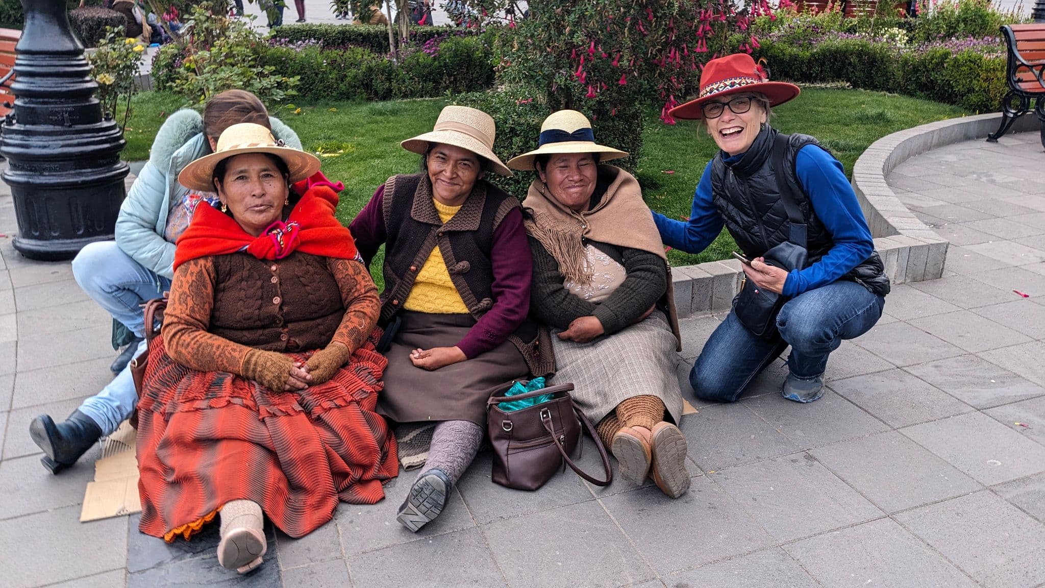 Three women in traditional Andean dress sitting on a plaza, posing with a tourist during a trip in Peru.