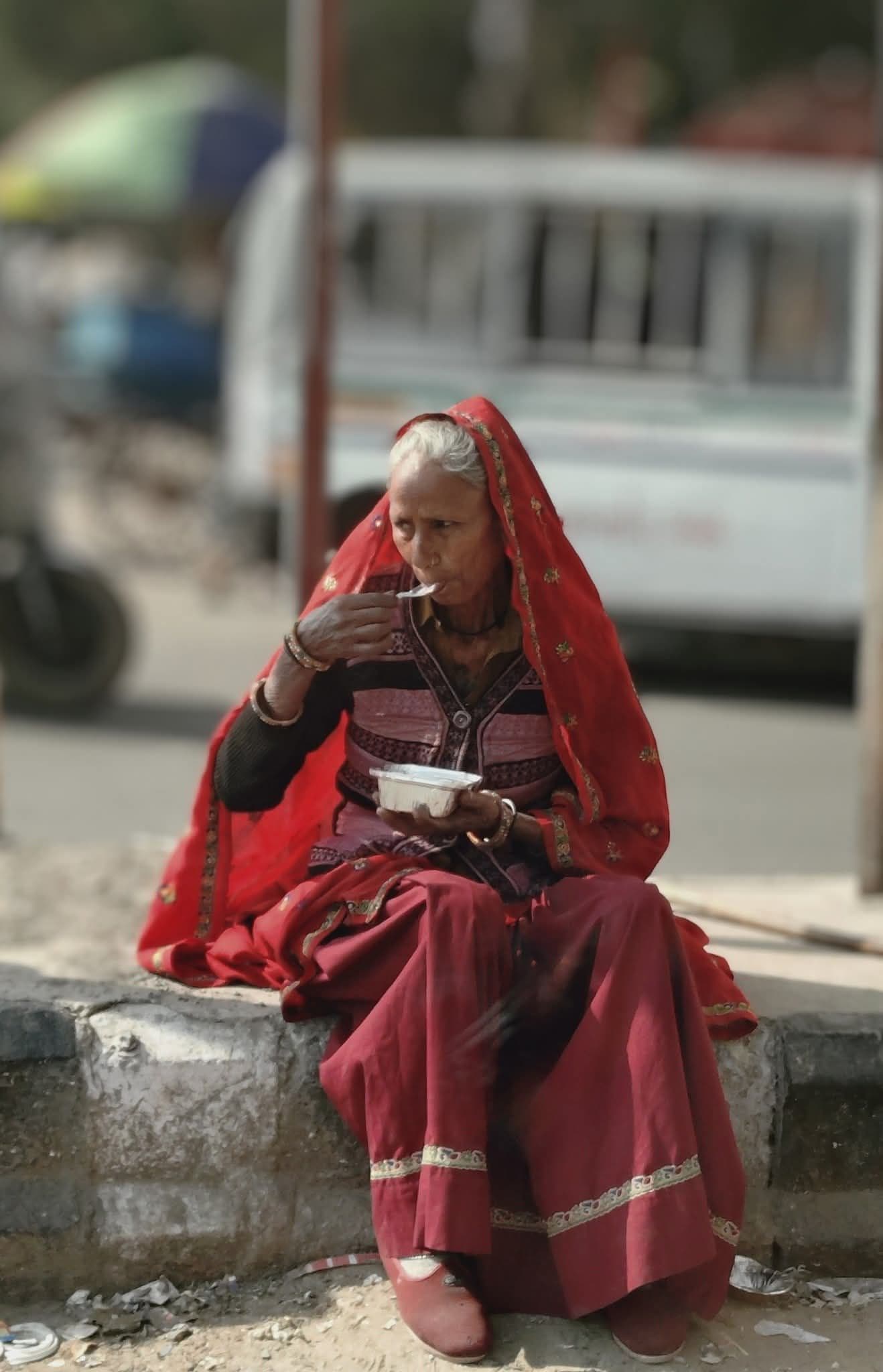 Elderly woman in a red sari eating from a takeaway container while seated on a street curb in Delhi, India.