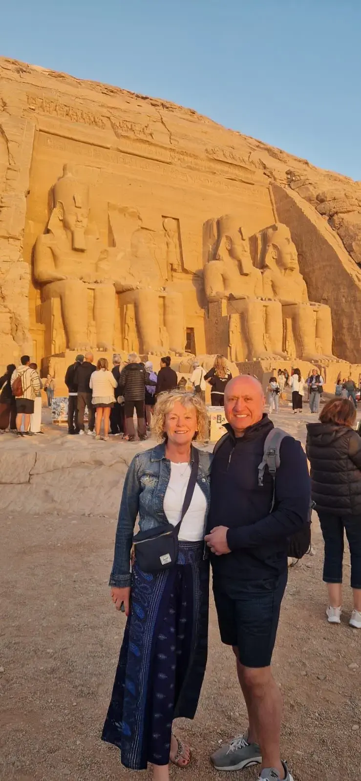 Abu Simbel temple in Egypt with two travelers posing in front of the colossal seated statues at sunset.