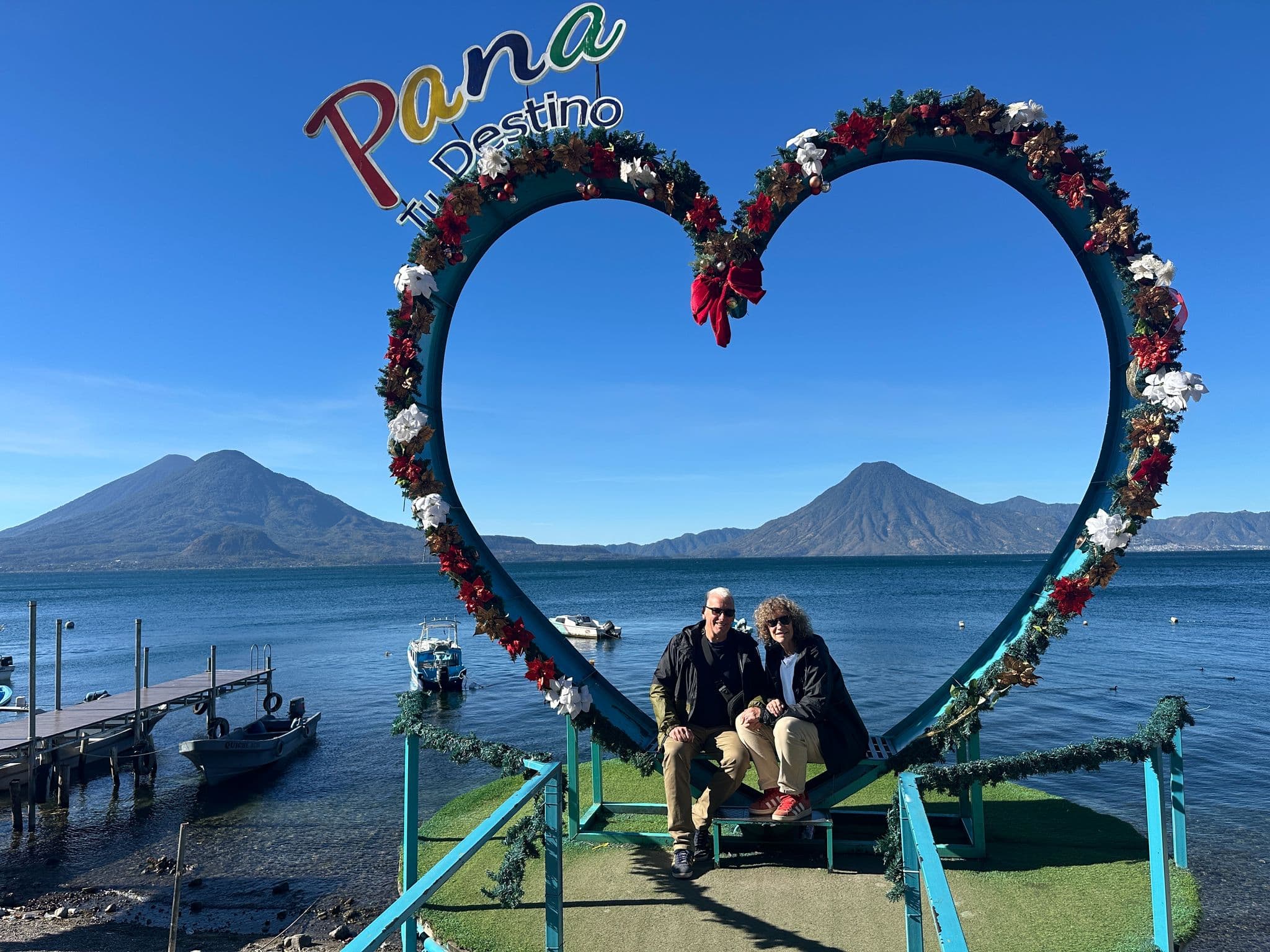 Lake Atitlán framed by a floral heart with two travelers seated on a platform, volcanoes rising behind, Panajachel, Guatemala.