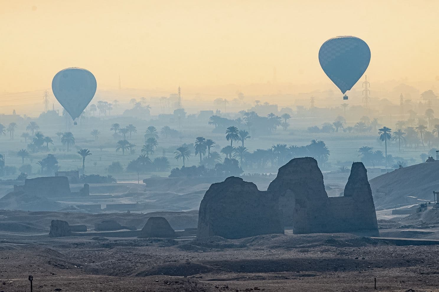 Two hot air balloons floating over ancient ruins and palm groves near Luxor, Egypt.