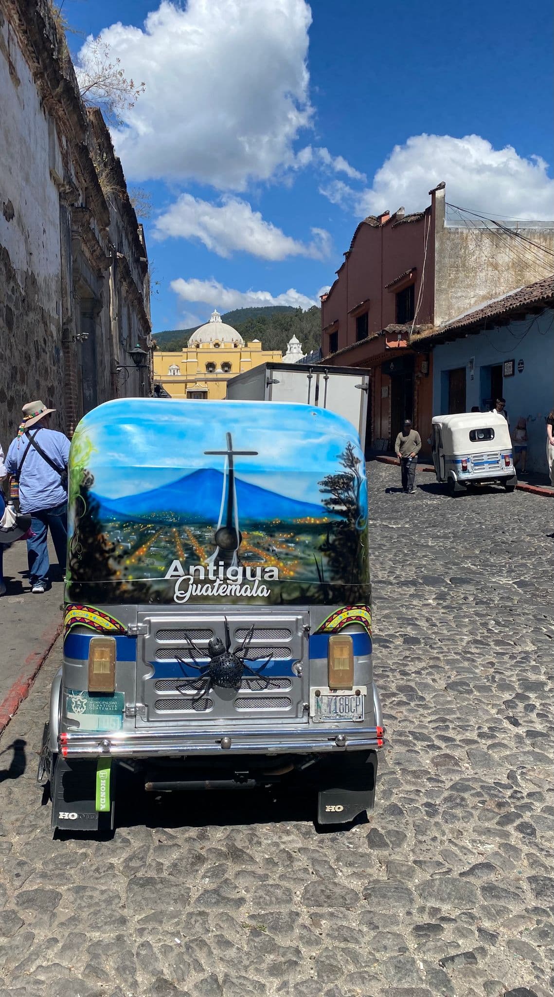Colorfully painted tuk-tuk parked on a cobblestone street with La Merced Church dome visible in Antigua, Guatemala.
