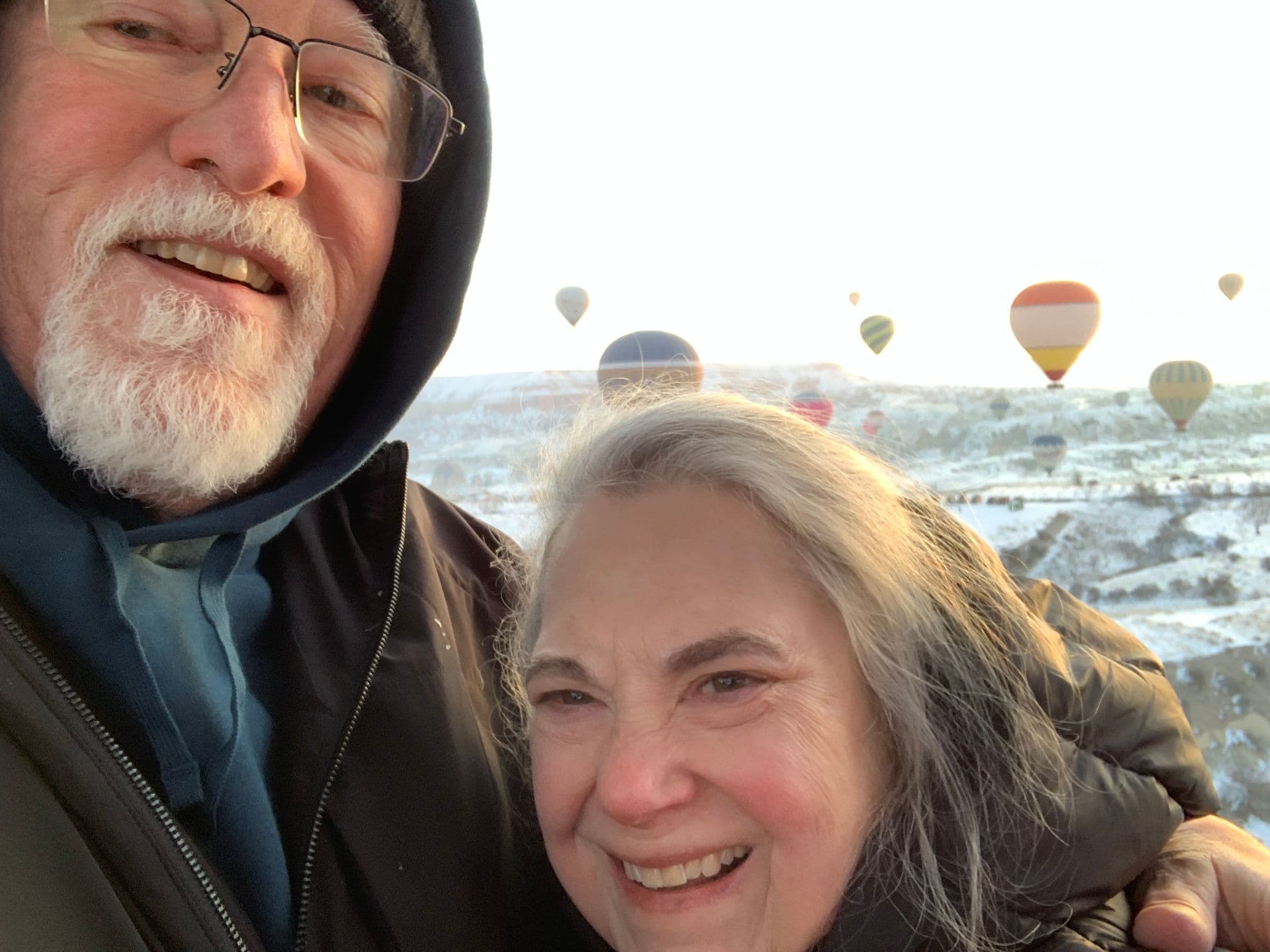 Hot air balloons over Cappadocia with two travelers smiling in a close selfie above a snowy valley, Turkey.
