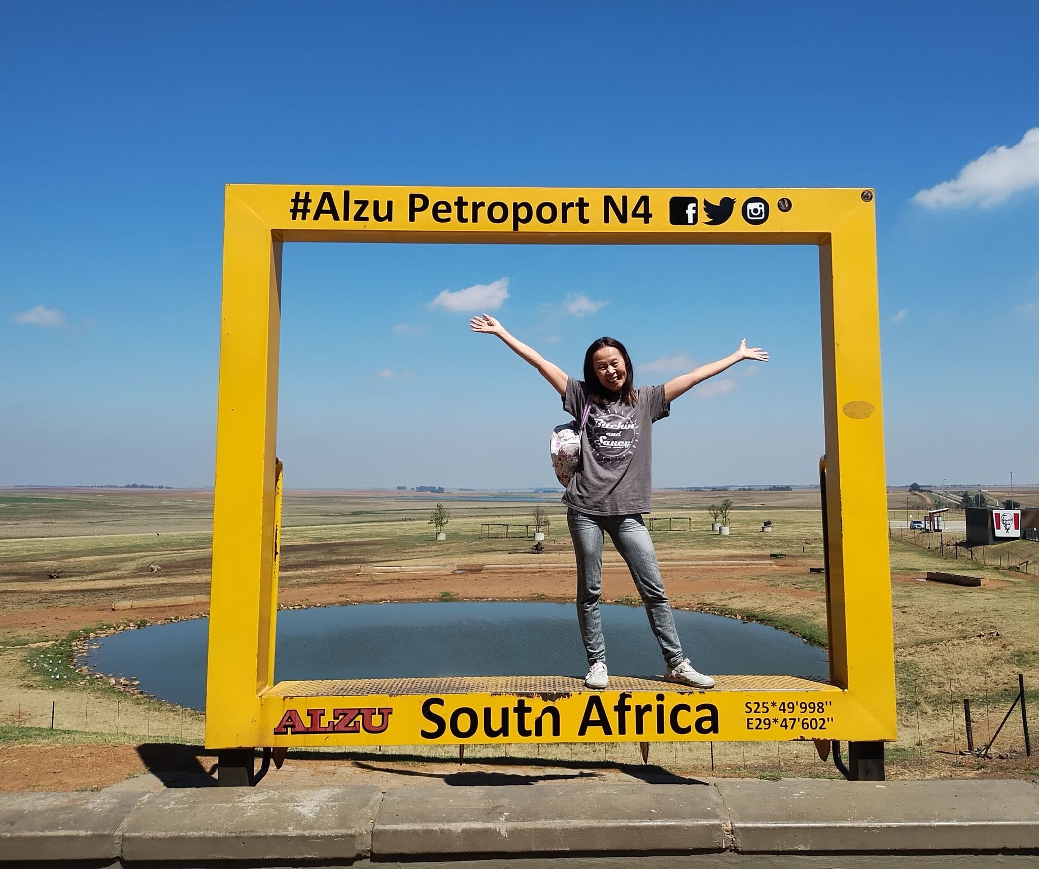 Yellow frame sign "Alzu Petroport N4" framing a woman with arms outstretched by a pond and open plains, South Africa.