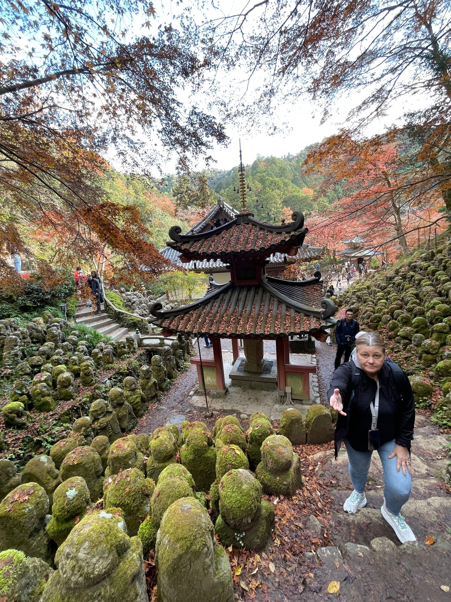 Otagi Nenbutsu-ji Temple's moss-covered rakan statues and pagoda, a traveler climbs stone steps reaching out, Arashiyama, Kyoto, Japan.