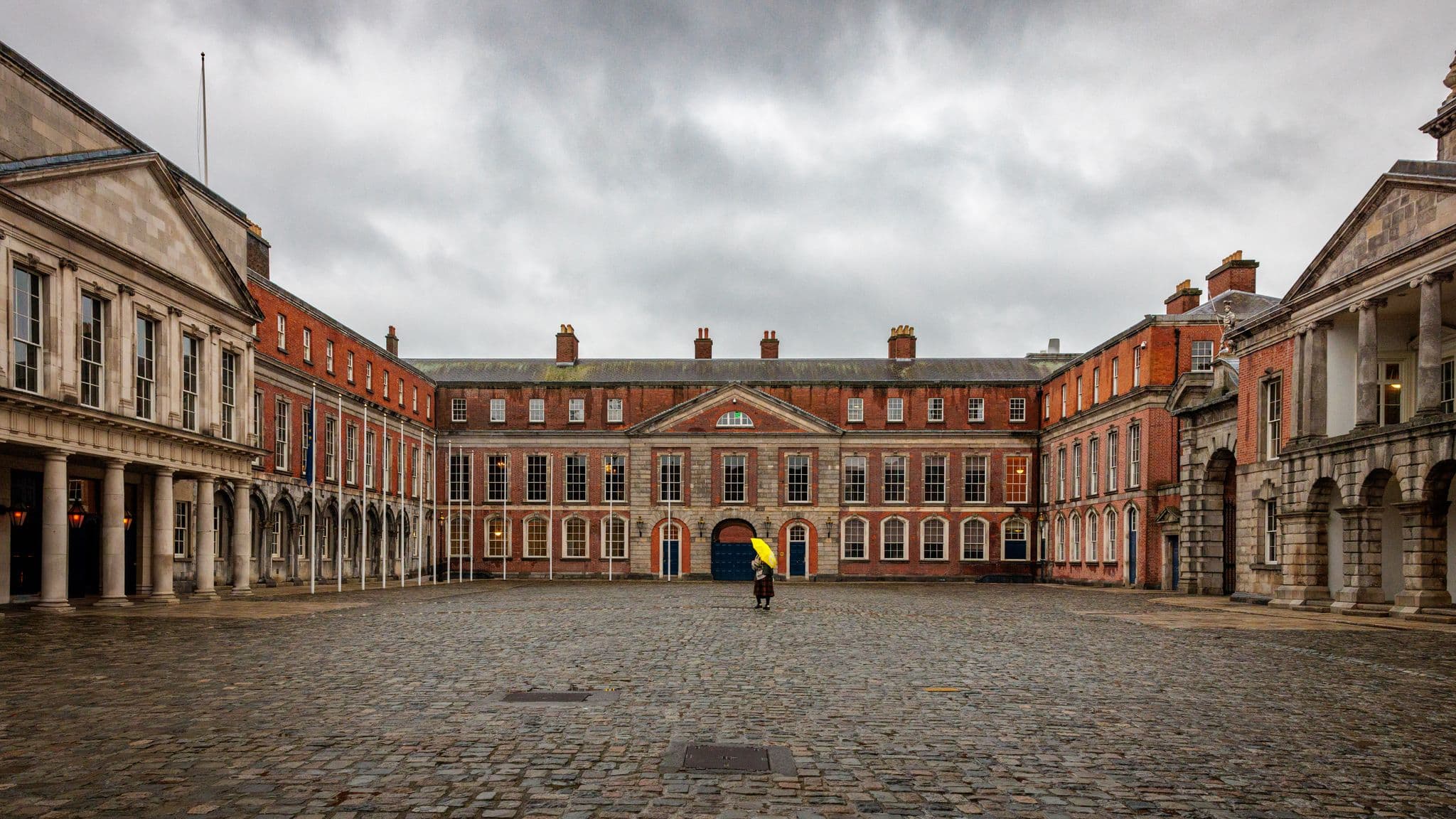 Dublin Castle courtyard with a person holding a yellow umbrella walking across wet cobblestones, Dublin, Ireland.